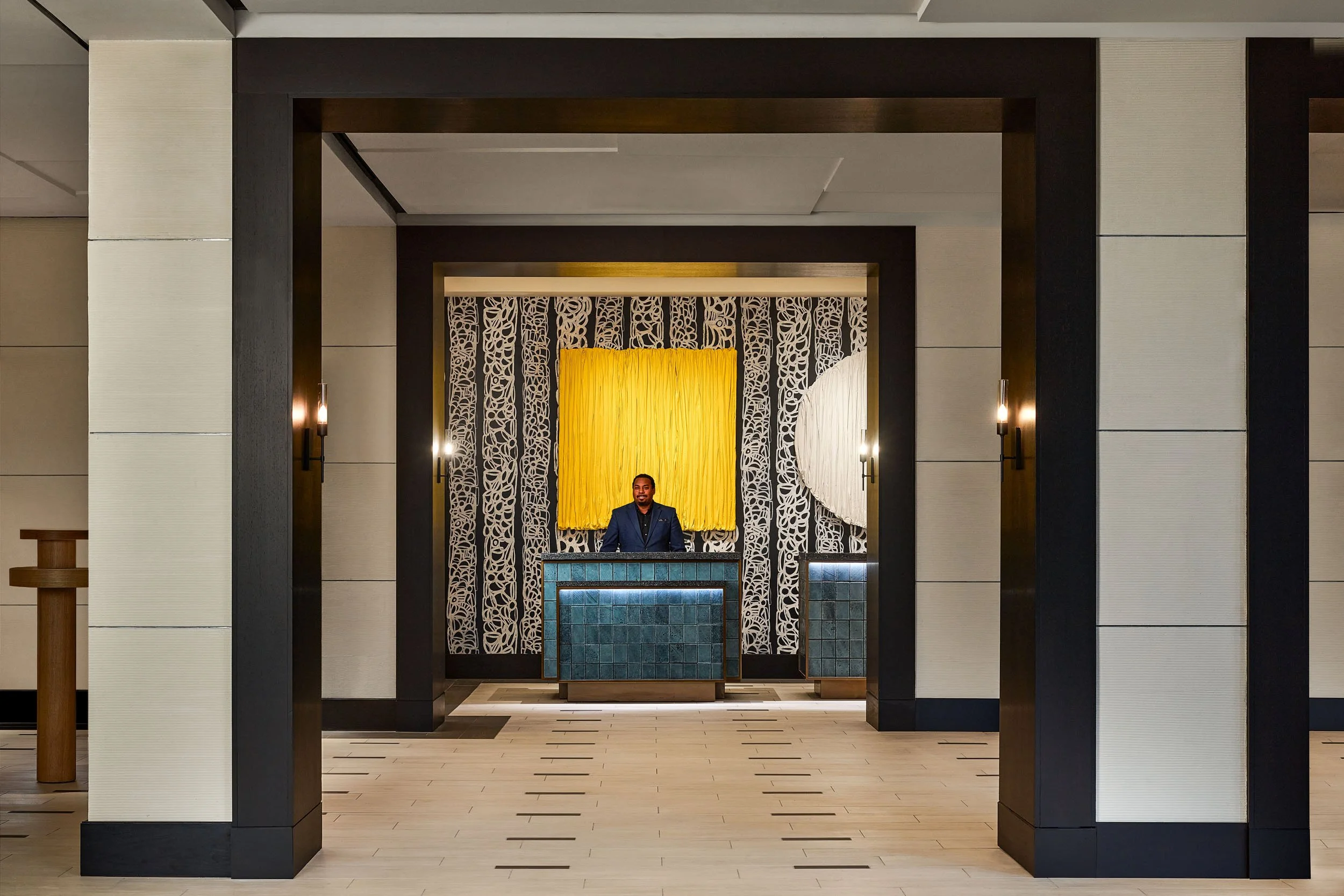 Interior of a hotel lobby with a reception desk, decorative black and white patterned panels, and a yellow wall hanging behind the reception area. A man in a dark suit stands at the desk.
