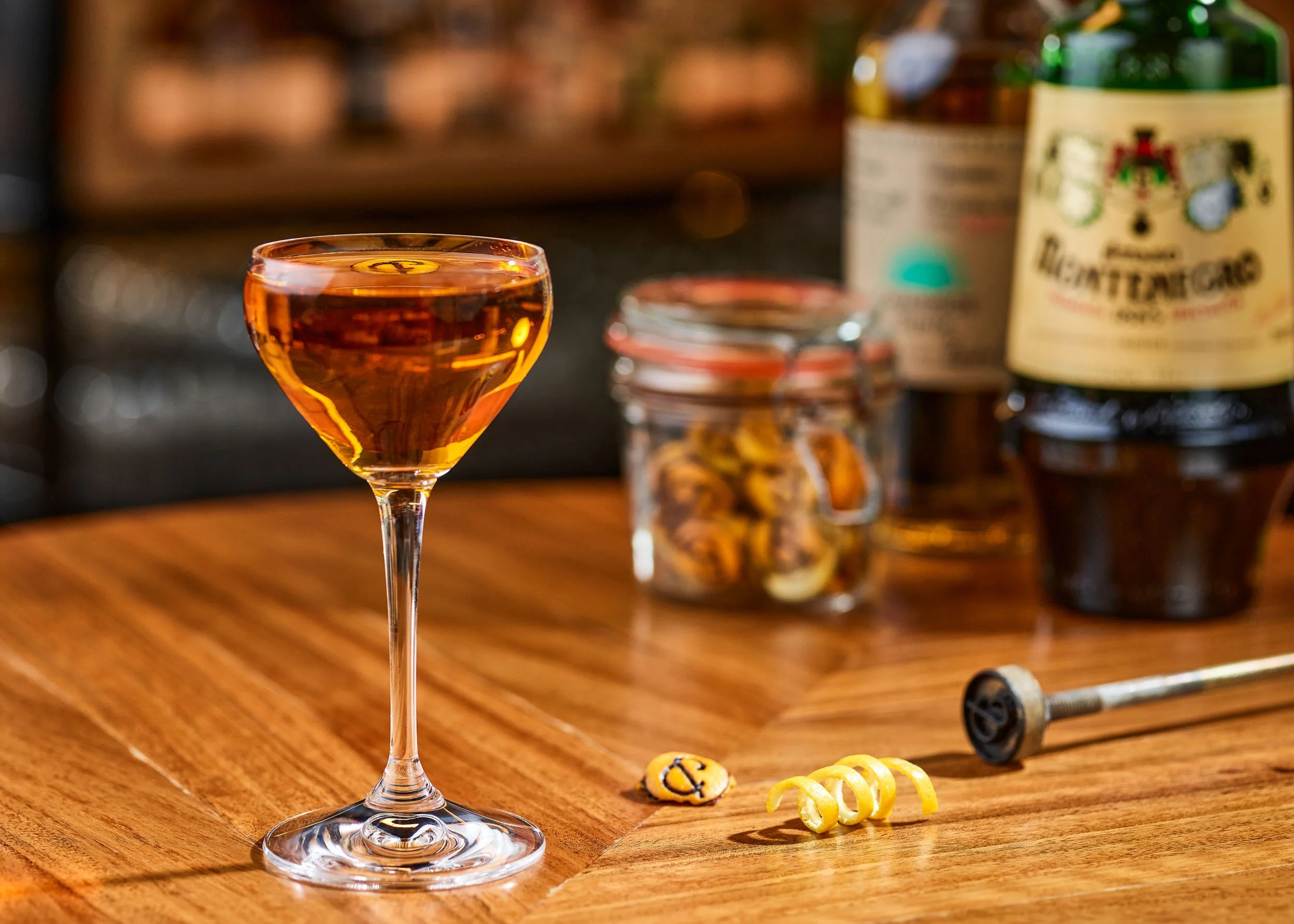 A cocktail glass filled with a dark amber drink on a wooden surface. In the background, there are candy-coated chocolates, a jar with cookies, and bottles of liquor.