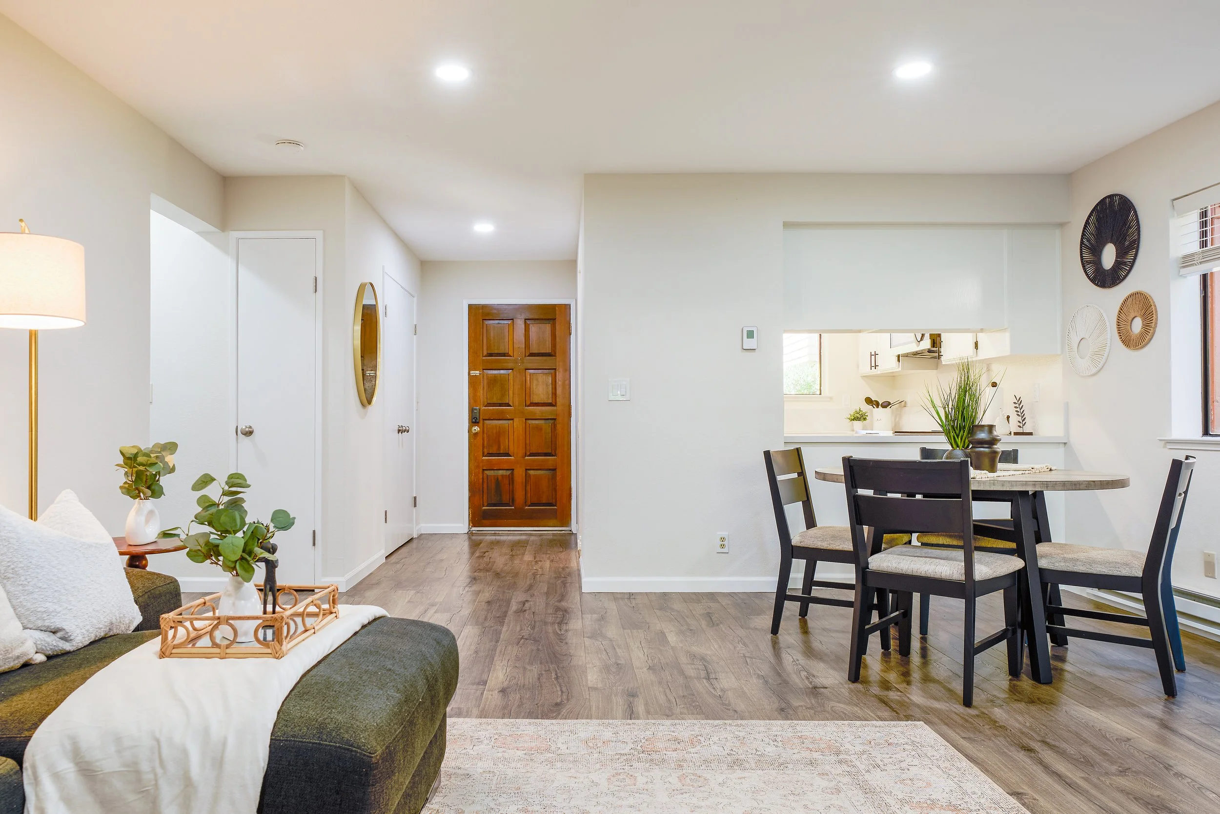 Living room with a gray sofa, white pillows, a small wooden tray, a plant in a white pot, and a lamp. The room has hardwood flooring, a front door, a dining area with a round table and black chairs, and wall decorations. Open kitchen area visible thr