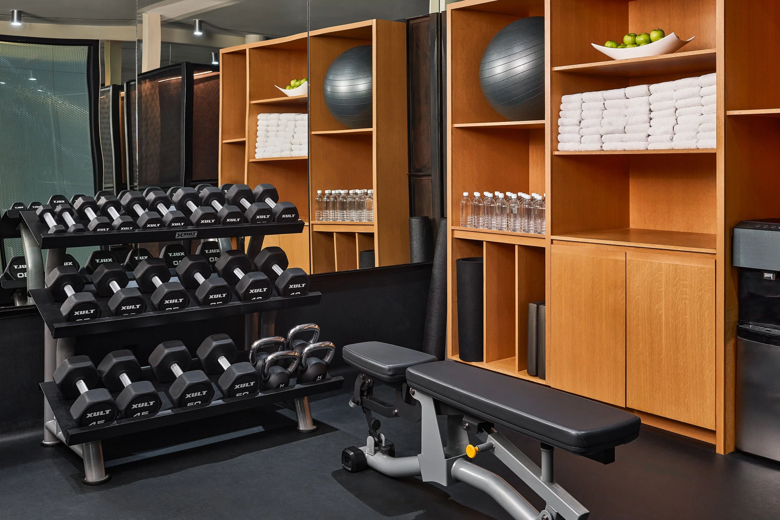 A gym locker room with a rack of black dumbbells, water bottles, and towels stored on wooden shelves. Fitness equipment and a bench are also visible.