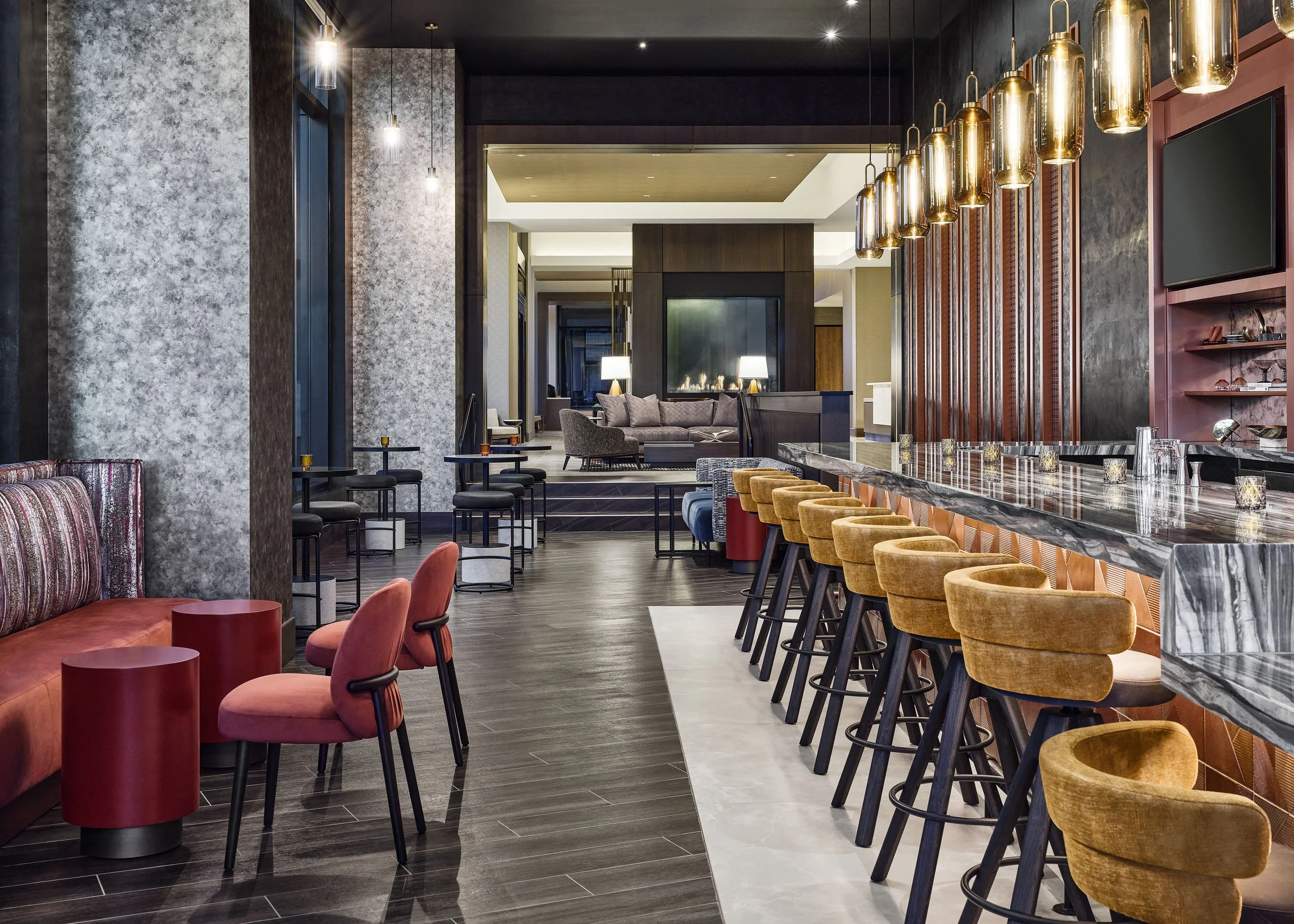 Modern hotel lobby with a marble-topped bar counter, mustard-colored bar stools, a sitting area with gray and purple furniture, and a fireplace with a wooden frame in the background. Gold pendant lights hang over the bar and a flat-screen TV is mount