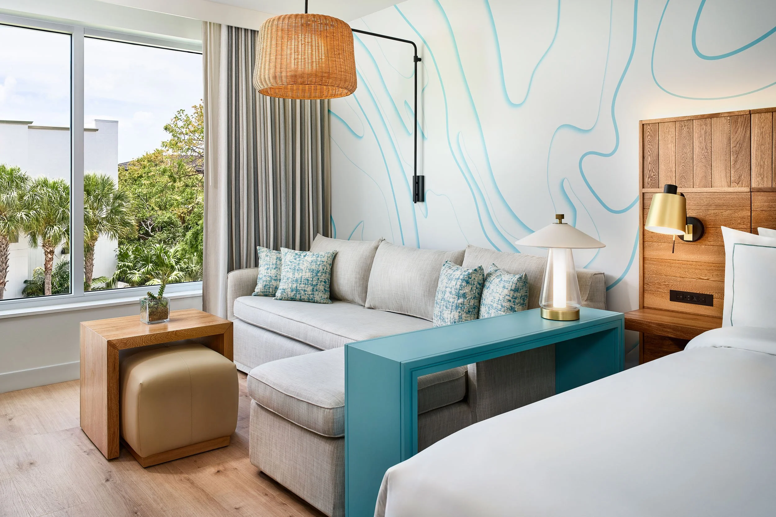 Modern hotel room with large window, beige sofa with blue and beige pillows, wooden side table with potted plant, wall art with blue lines, white lamp with gold base, and partial view of bed with white bedding.