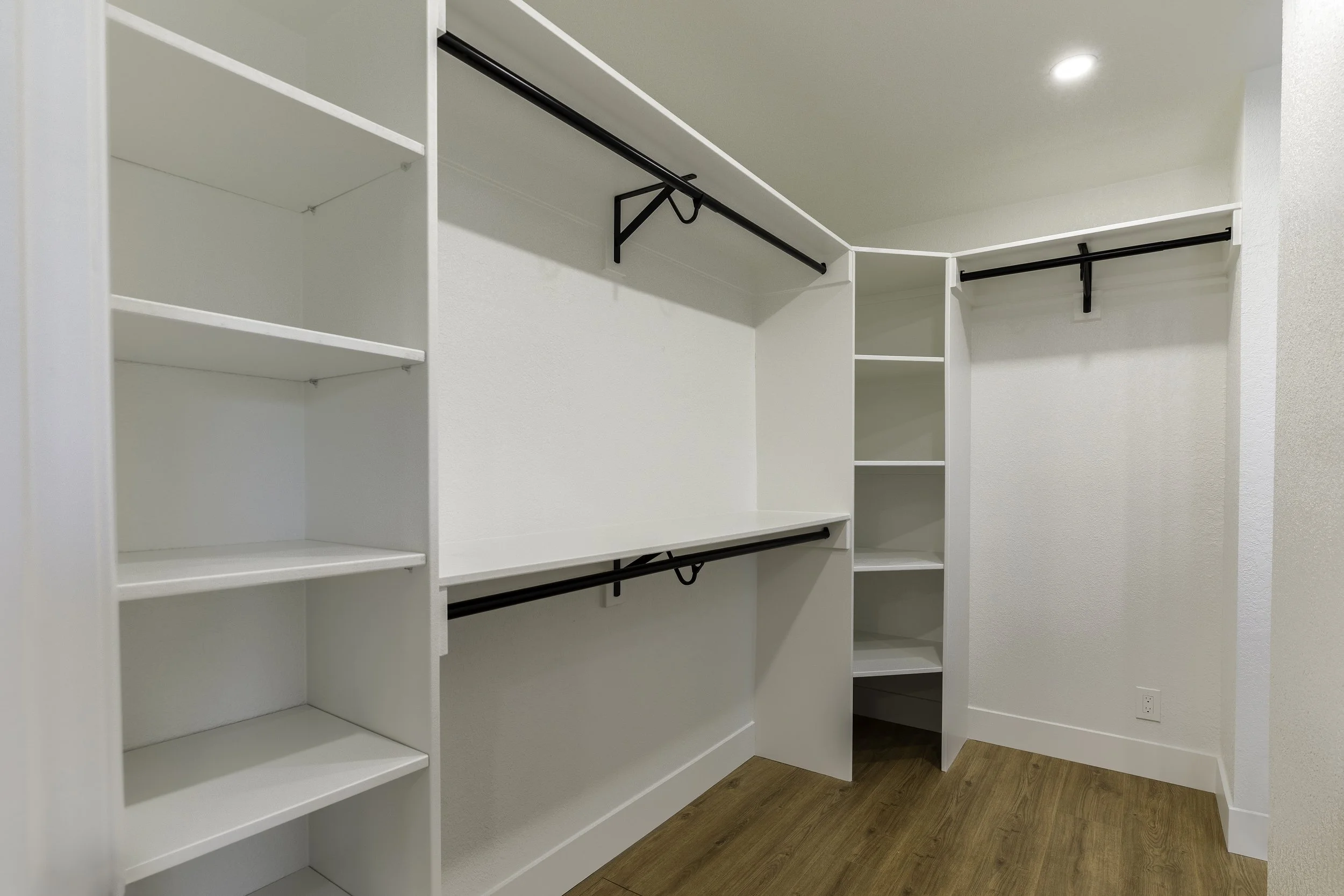 Empty white closet with multiple shelves and hanging rods, wooden floor, and a ceiling light.