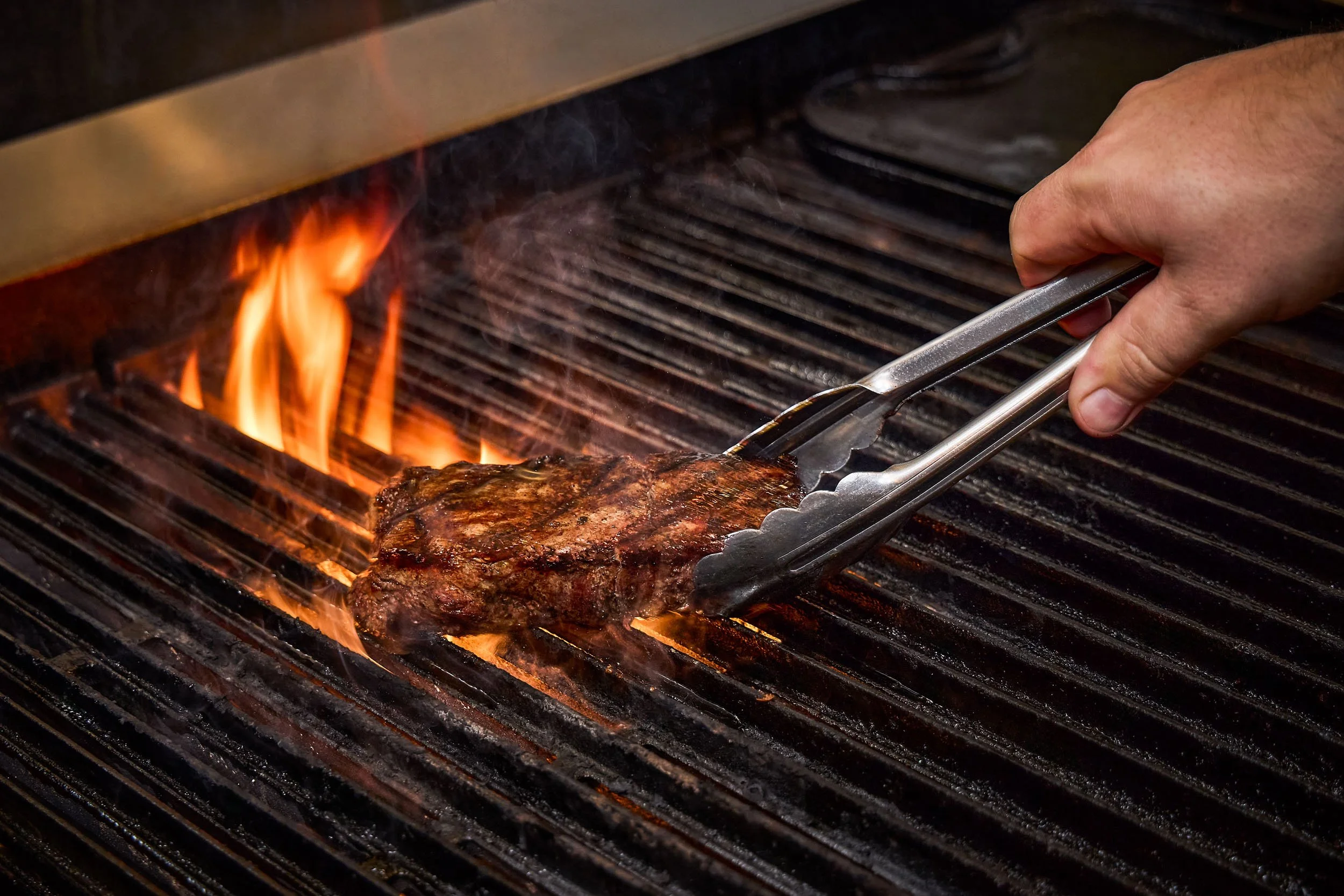 A close-up of a hand using tongs to grill a cooked steak on a barbecue grill with visible flames and heat lines.