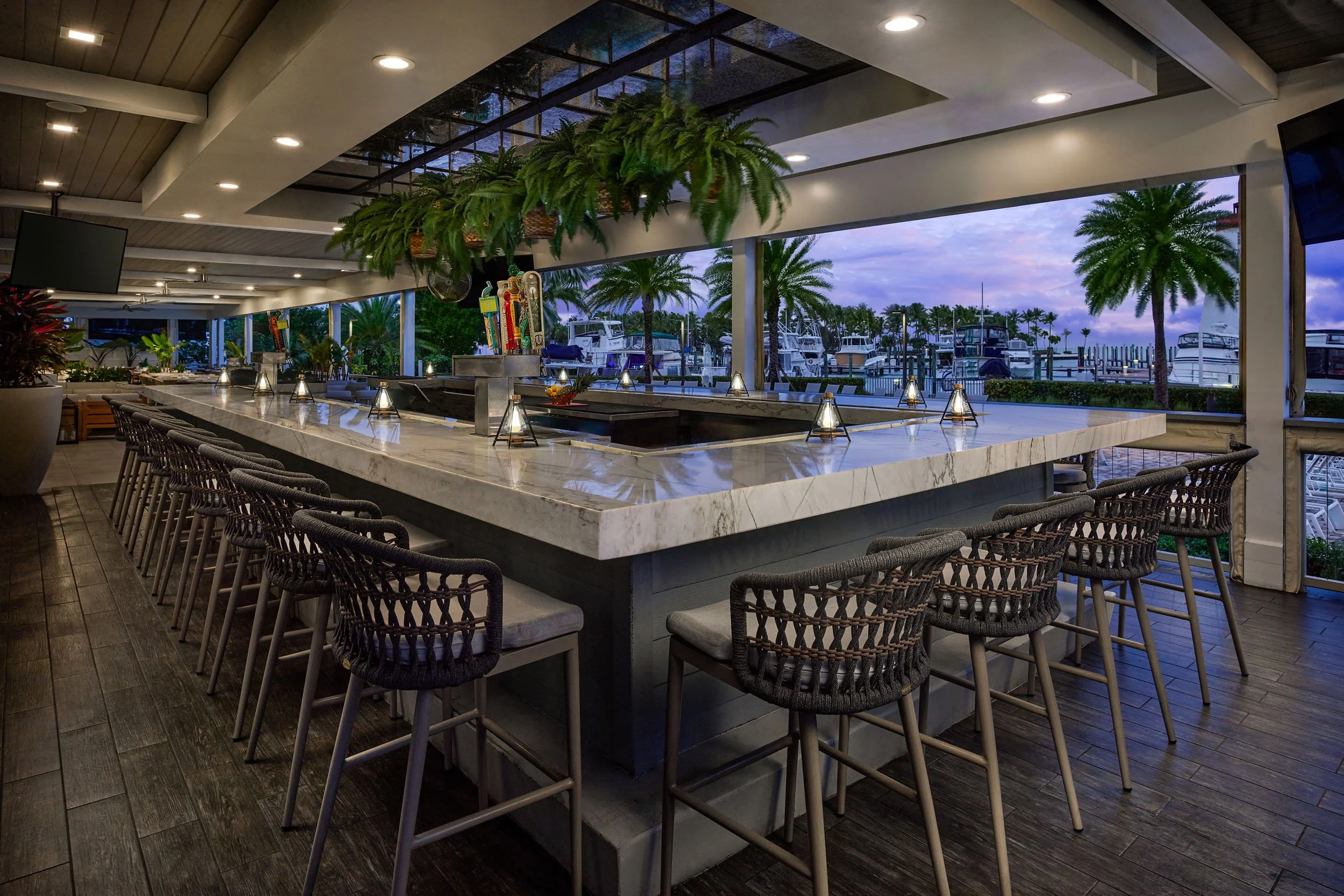 Indoor bar area overlooking boats docked at a marina with palm trees outside, evening sky, marble bar counter, chairs, hanging plants, and television screens.