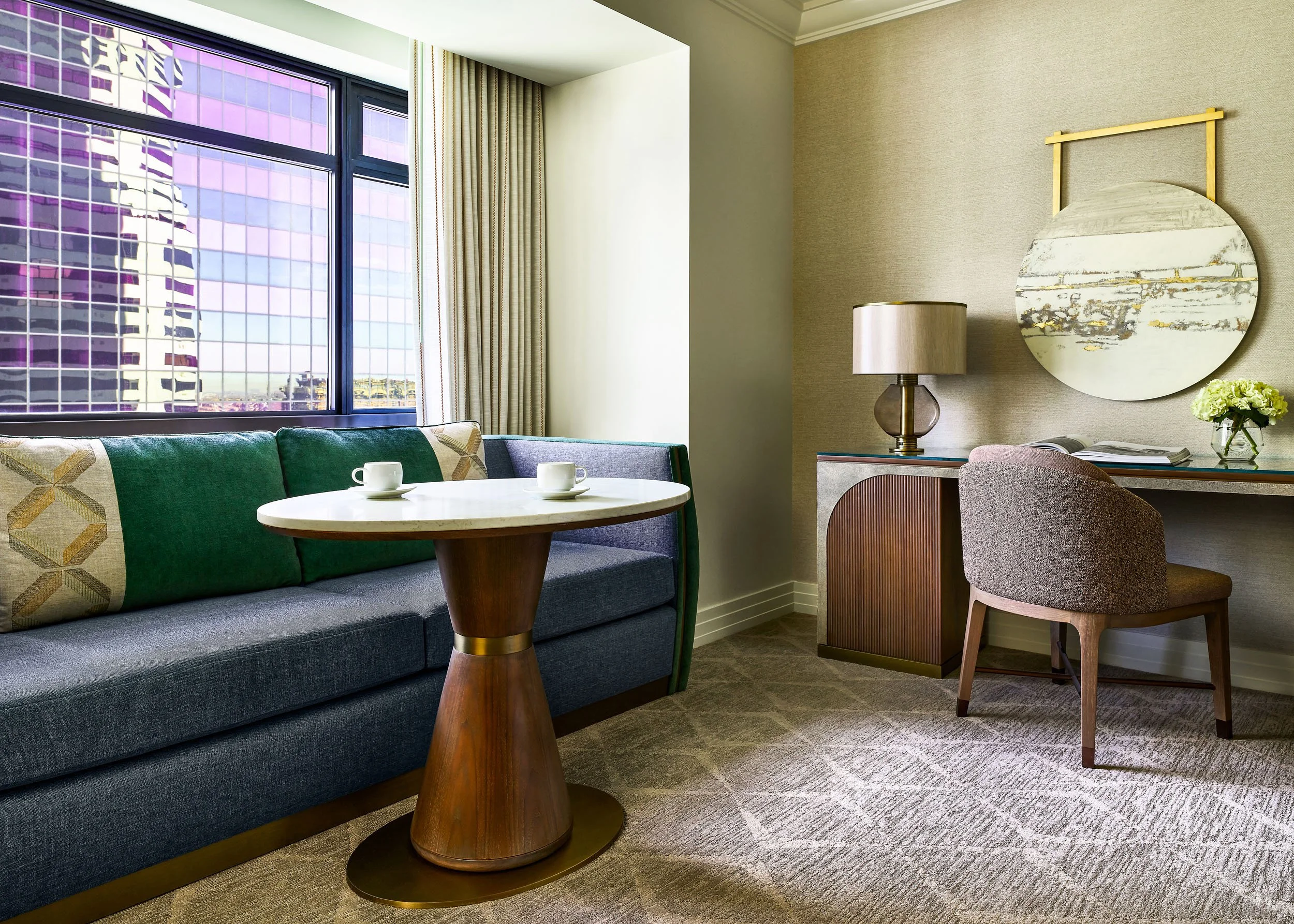Interior of a modern apartment living room, featuring a large window with city views, a gray sofa with geometric patterned cushions, a round wooden coffee table with two white cups, a table lamp with a silver base, abstract wall art with circular mir