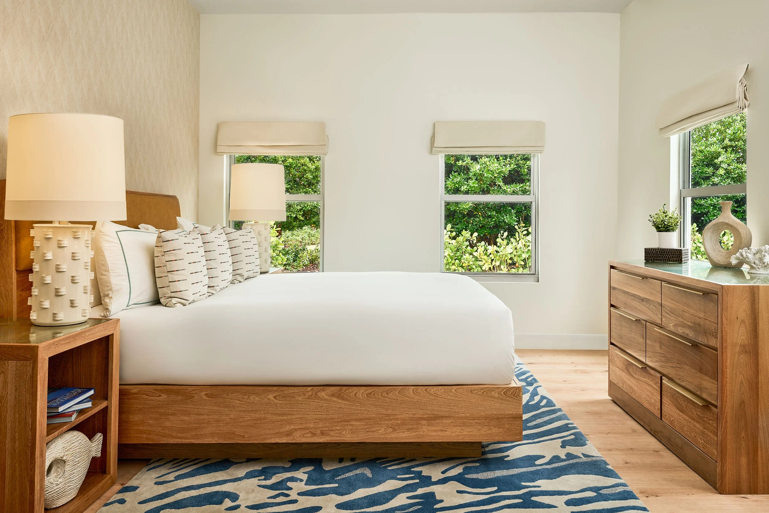 A bedroom with a white bed, wooden bedside table with books and a lamp, wooden dresser with decor, and three windows showing green foliage outside.