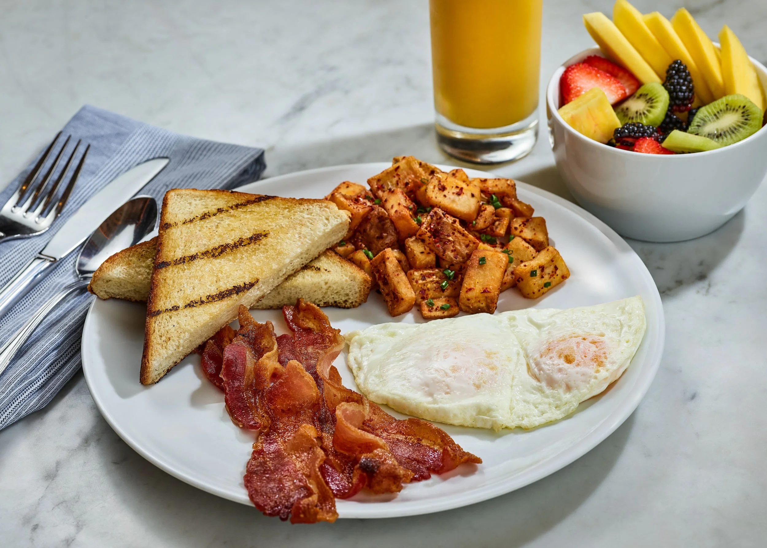 A breakfast plate with toast, scrambled eggs, bacon, and spicy potatoes, served with a side of mixed fresh fruit, and a glass of orange juice on a marble table with a fork, knife, and napkin.