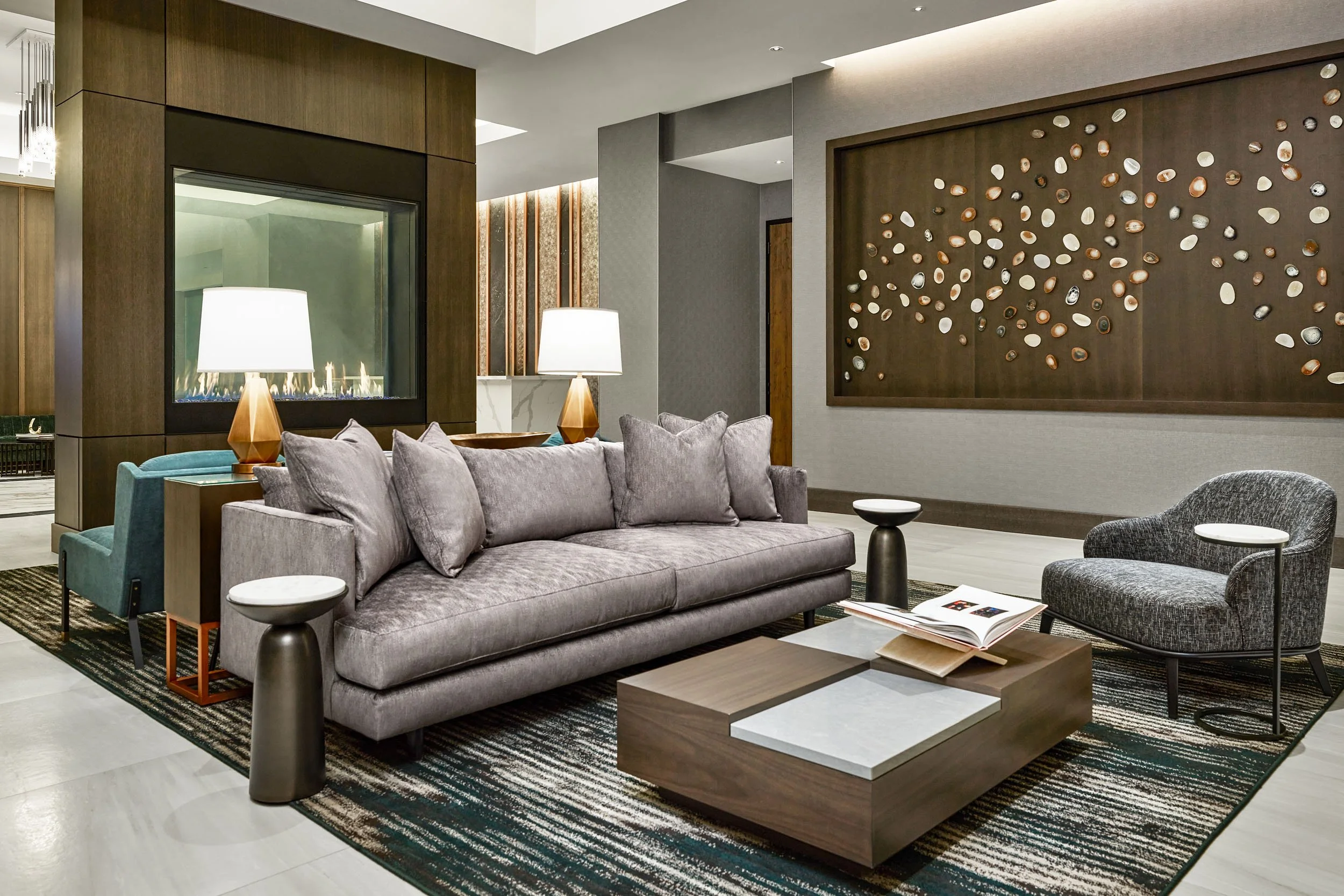 Modern hotel lobby featuring a gray sofa with plush pillows, two wooden side tables with lamps, a geometric coffee table, and a large wall art piece with metallic shells, with a fireplace and decorative vertical lights in the background.