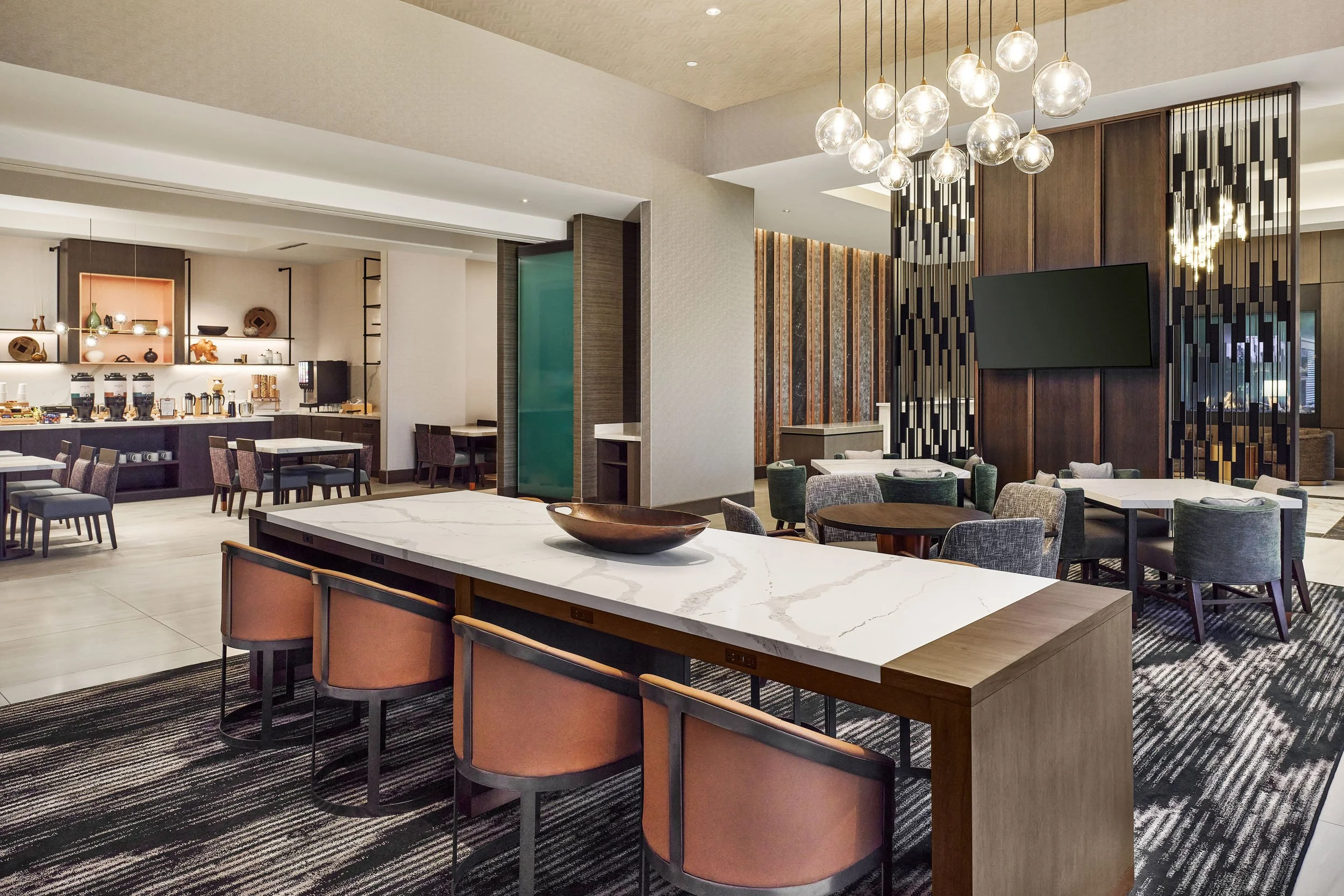 Modern hotel or restaurant dining area with a white marble island and pink cushioned chairs, dark wood tables, a coffee or refreshment station with coffee dispensers, and decorative lighting fixtures, including a chandelier with multiple glass globes