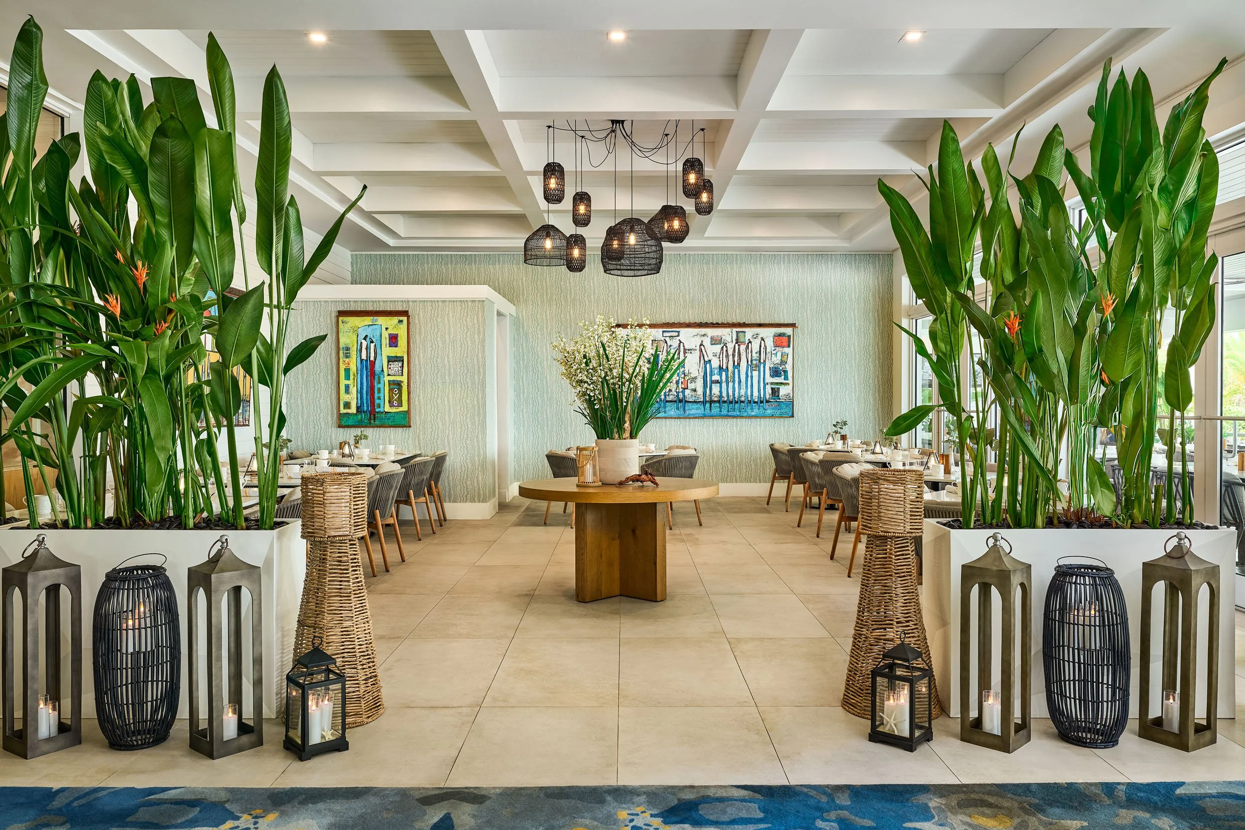 A spacious, modern indoor dining area with tropical plants and decorative lanterns, featuring a central table with a floral arrangement and colorful artwork on the walls.