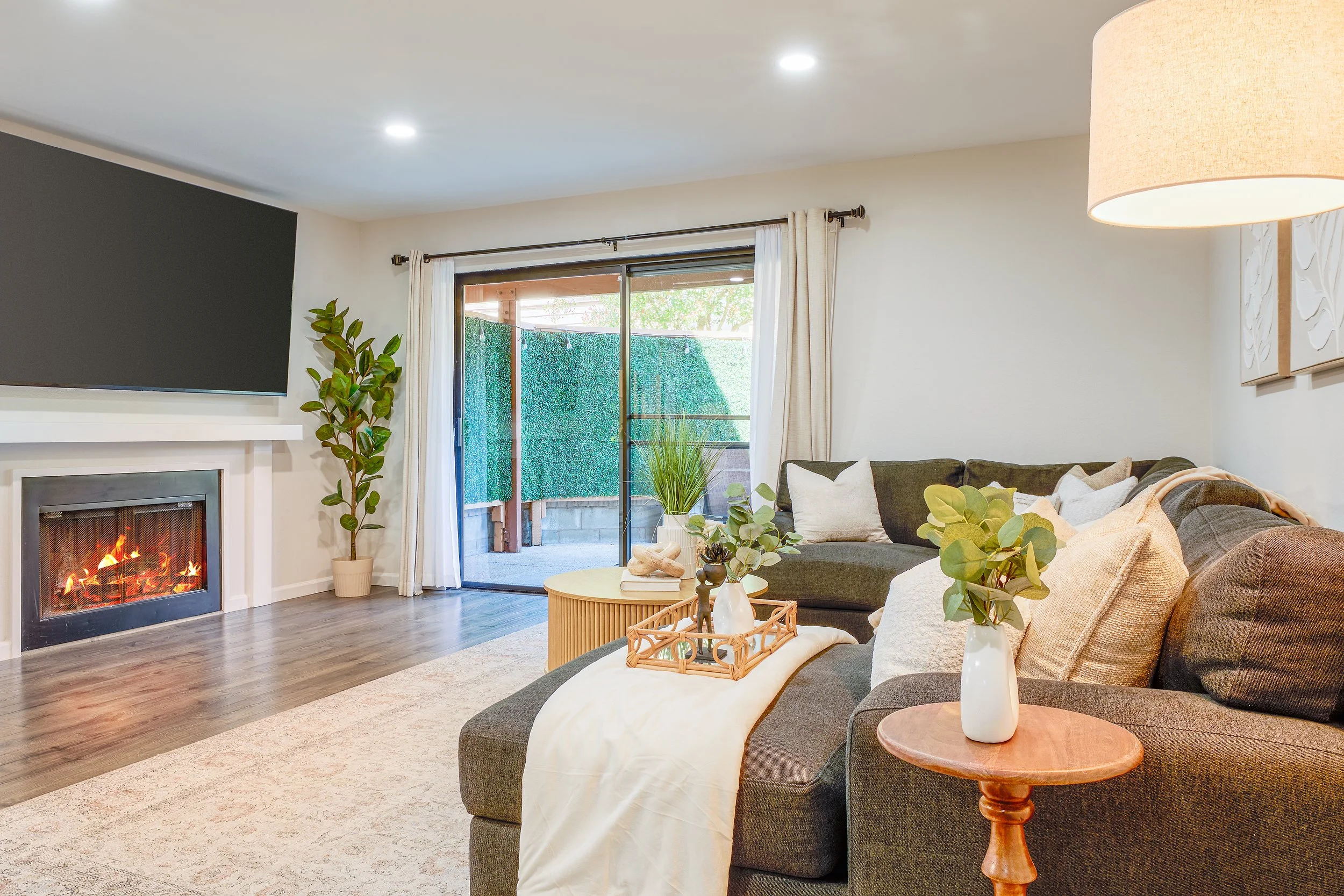 Living room with a fireplace, a large wall-mounted TV, a sliding glass door leading to an outdoor area, a sectional sofa with pillows, a wooden side table with a white lamp, plants, and decorative items.