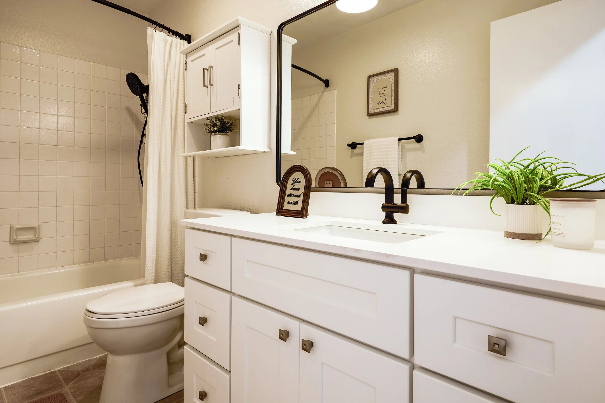 A bathroom with a white vanity, black faucet, and matching black handles. There is a mirror above the sink, a plant, and a framed sign on the vanity that reads, 'You have entered a calm zone.' The bathroom includes a shower with a white curtain, beig