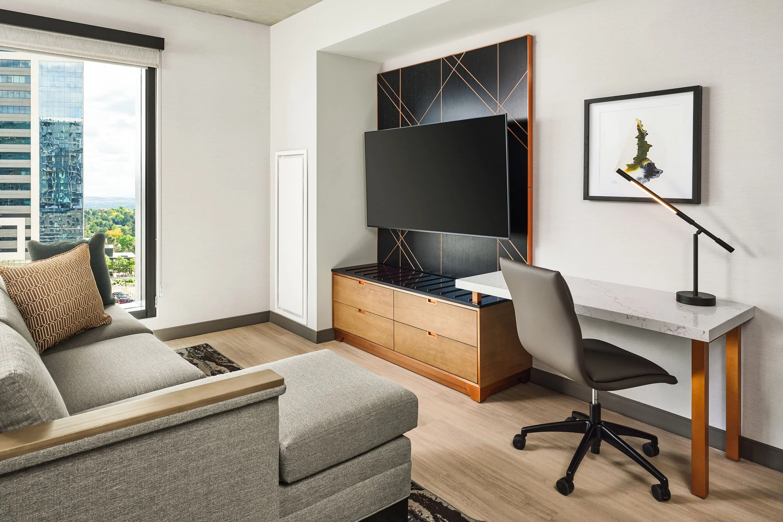 Modern living room with sofa, window view of city skyline and office space with desk, chair, and artwork.