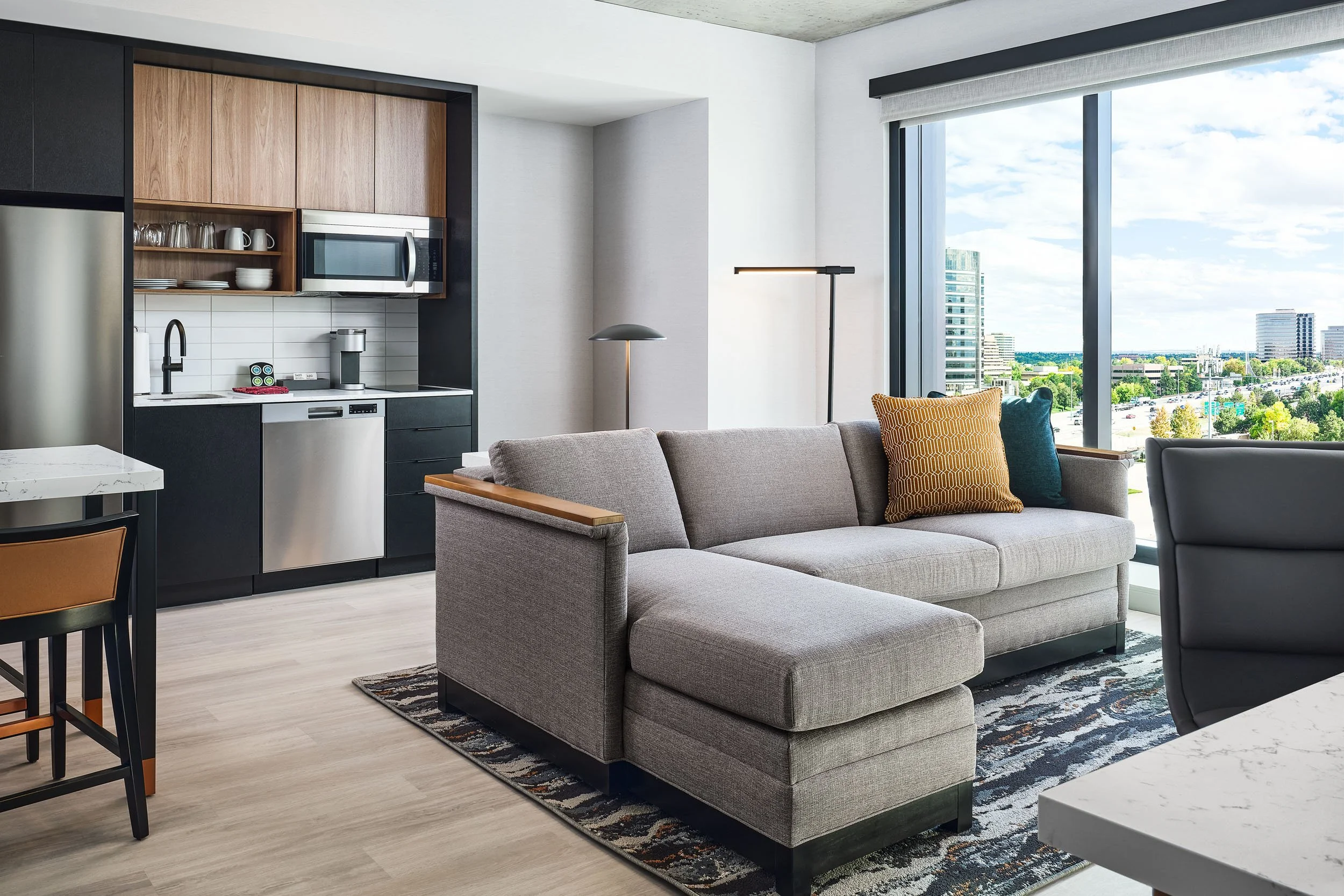 Modern living room with a gray sofa with yellow and blue pillows, a patterned rug, a large window showing a city view, and a kitchen area with wooden and black cabinets, a microwave, and a white backsplash.