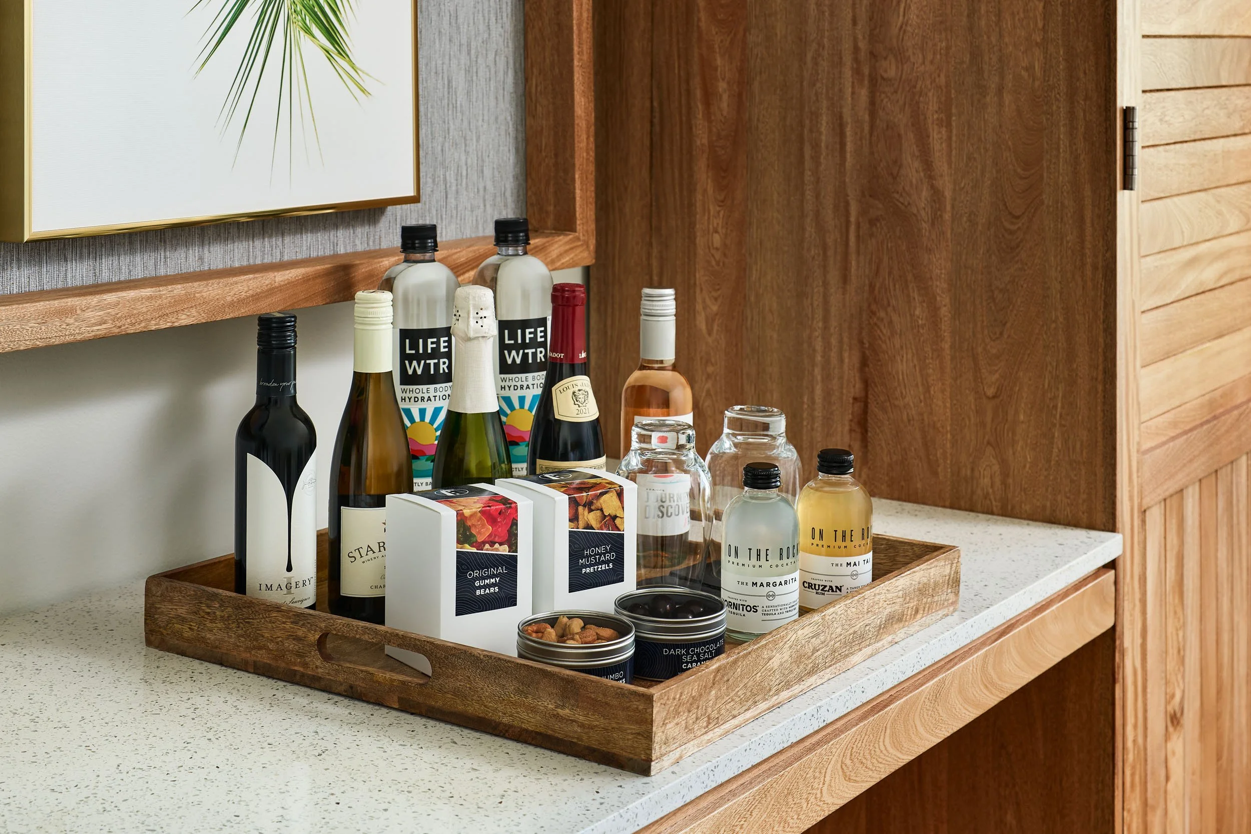 Wooden tray on a white countertop holding various bottles, boxes, and jars of snacks and beverages in a kitchen setting.