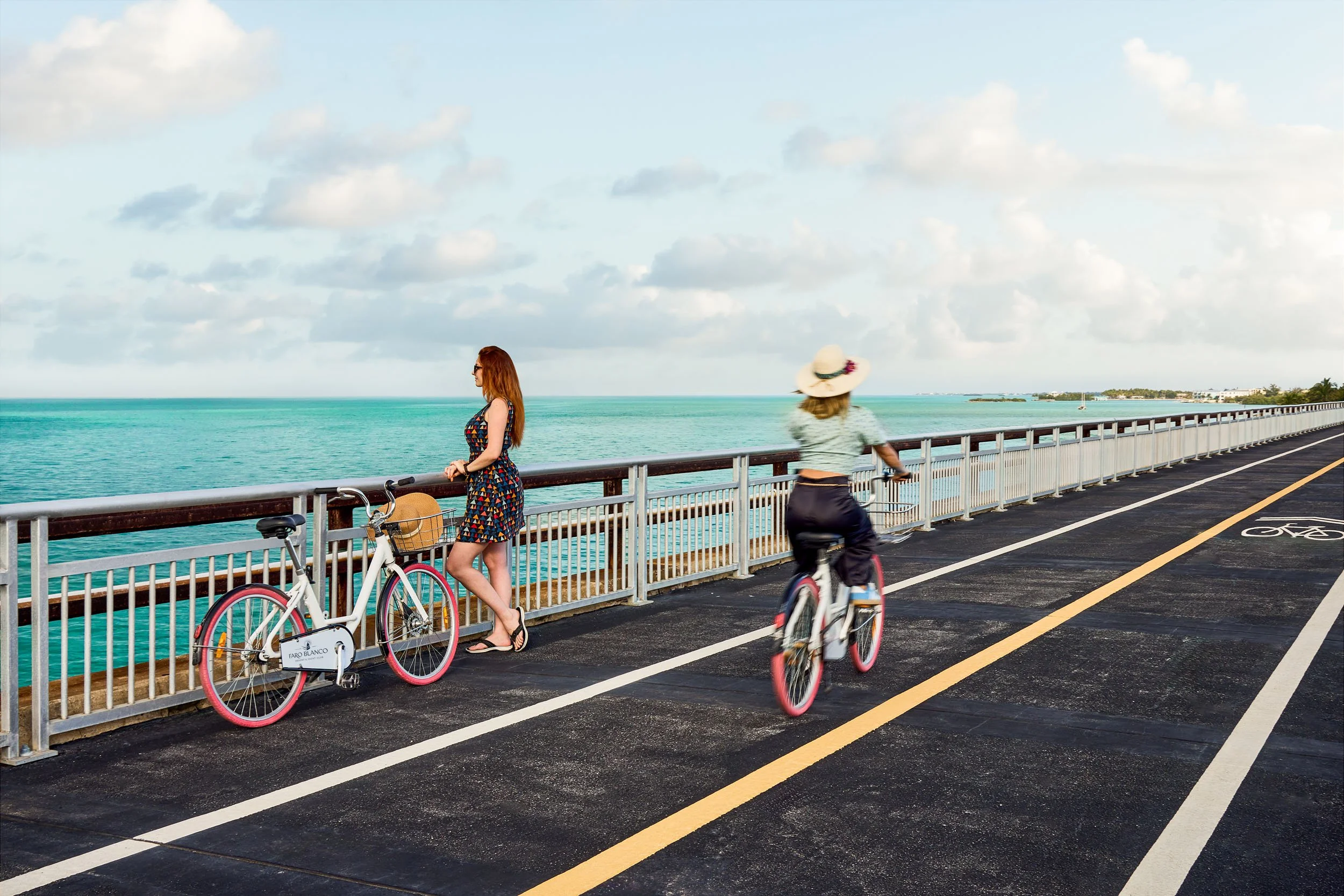 Two women with bicycles on a seaside bike path, one standing and the other riding, with a railing overlooking the ocean and partly cloudy sky in the background.