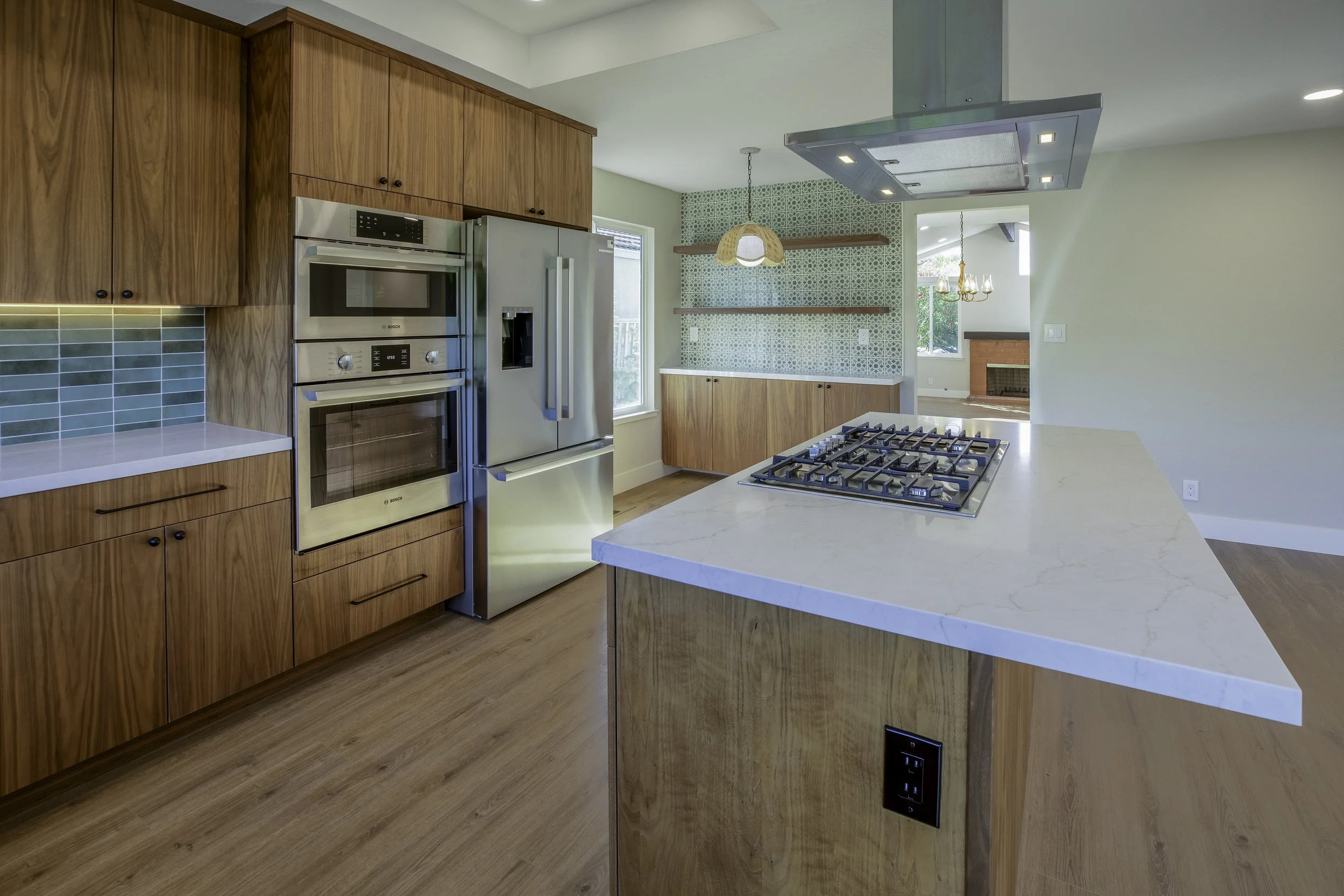 Modern kitchen with wooden cabinets, stainless steel appliances, a white marble island with a gas stove, a tiled backsplash, and a view into a dining room with a chandelier and a fireplace.