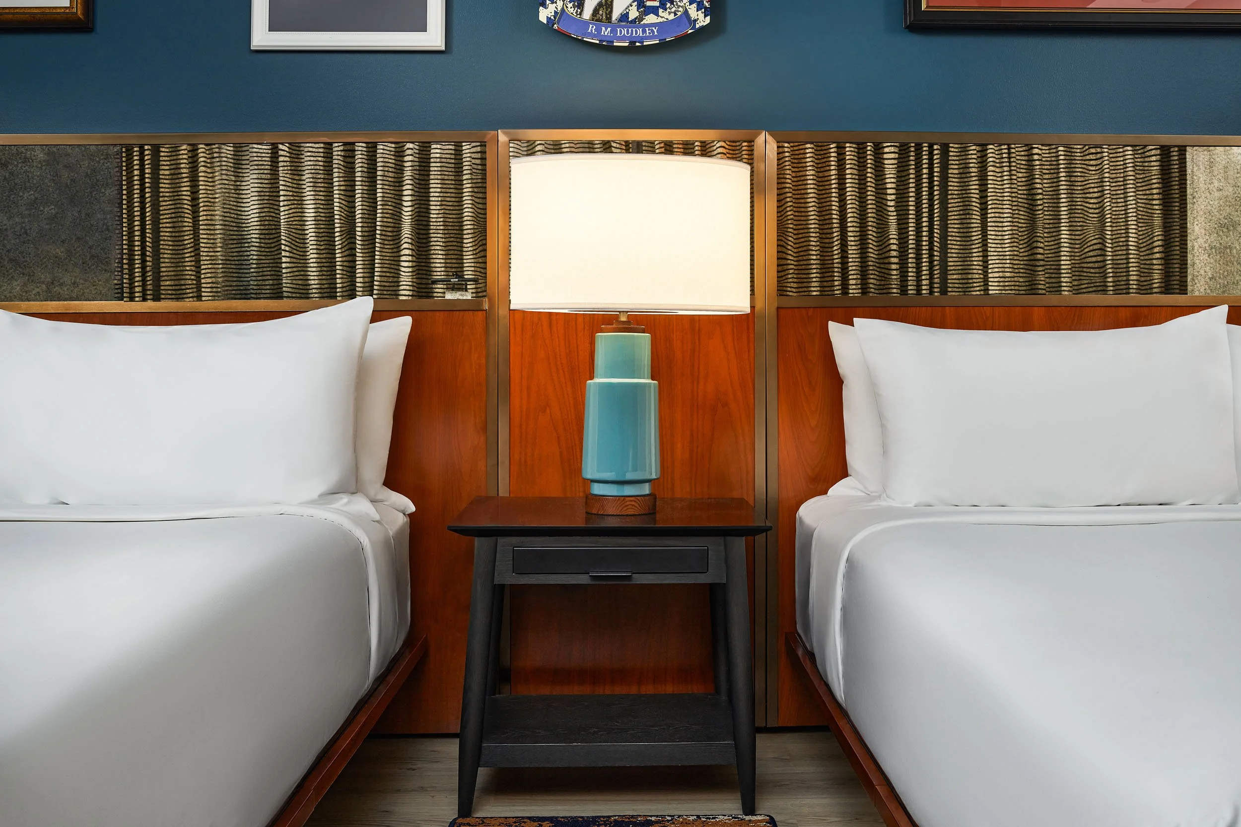 Two beds in a hotel room with white linens and wooden headboards separated by a small bedside table. A turquoise lamp is on the table and a wall with framed pictures and a blue wall in the background.