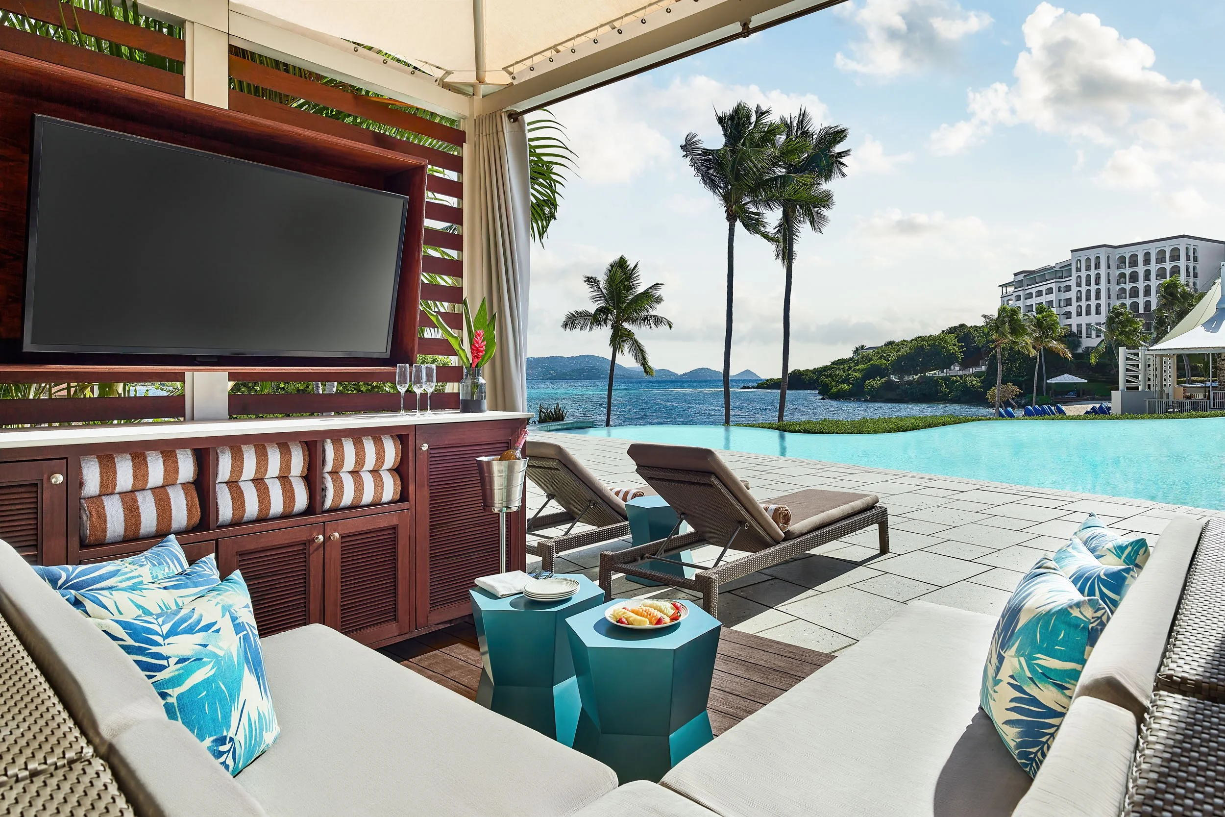 An outdoor poolside lounge area with chairs, blue and white tropical pillows, a small table with a bowl of fruit, and a view of palm trees, a building, and the ocean in the background.
