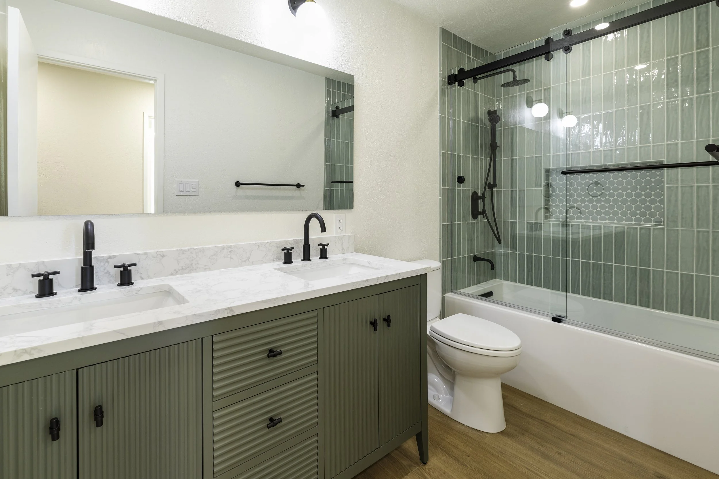 Modern bathroom with green cabinet vanity, white sink with black fixtures, large mirror, wood flooring, and a shower with green tiles, black fixtures, and a sliding glass door.