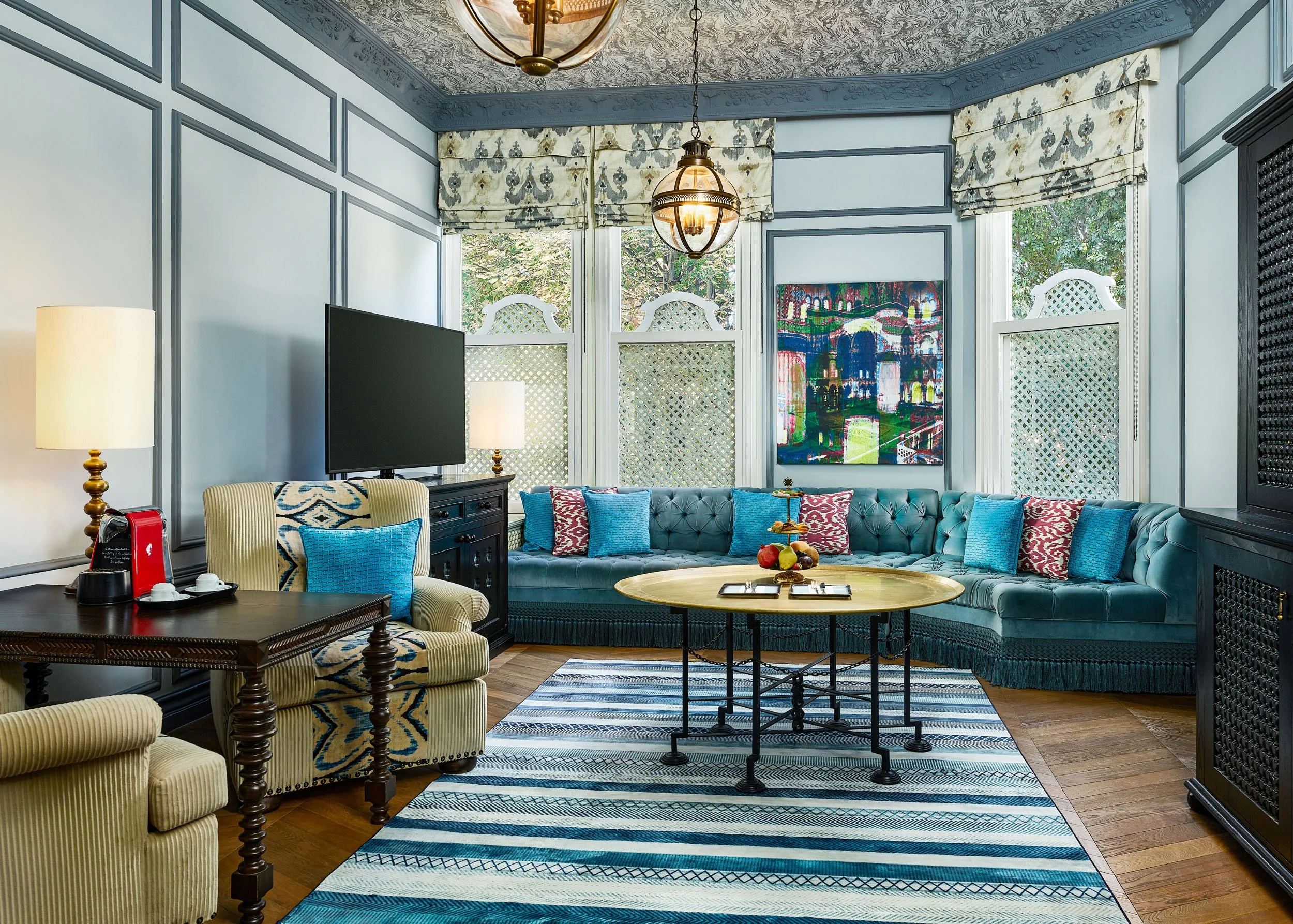 A living room with blue walls, crown molding, a large gray tufted sofa with red and blue pillows, a blue and white striped rug, a rectangle glass-top coffee table, a black sideboard, a flat-screen TV, a lamp with a white shade, and large windows with