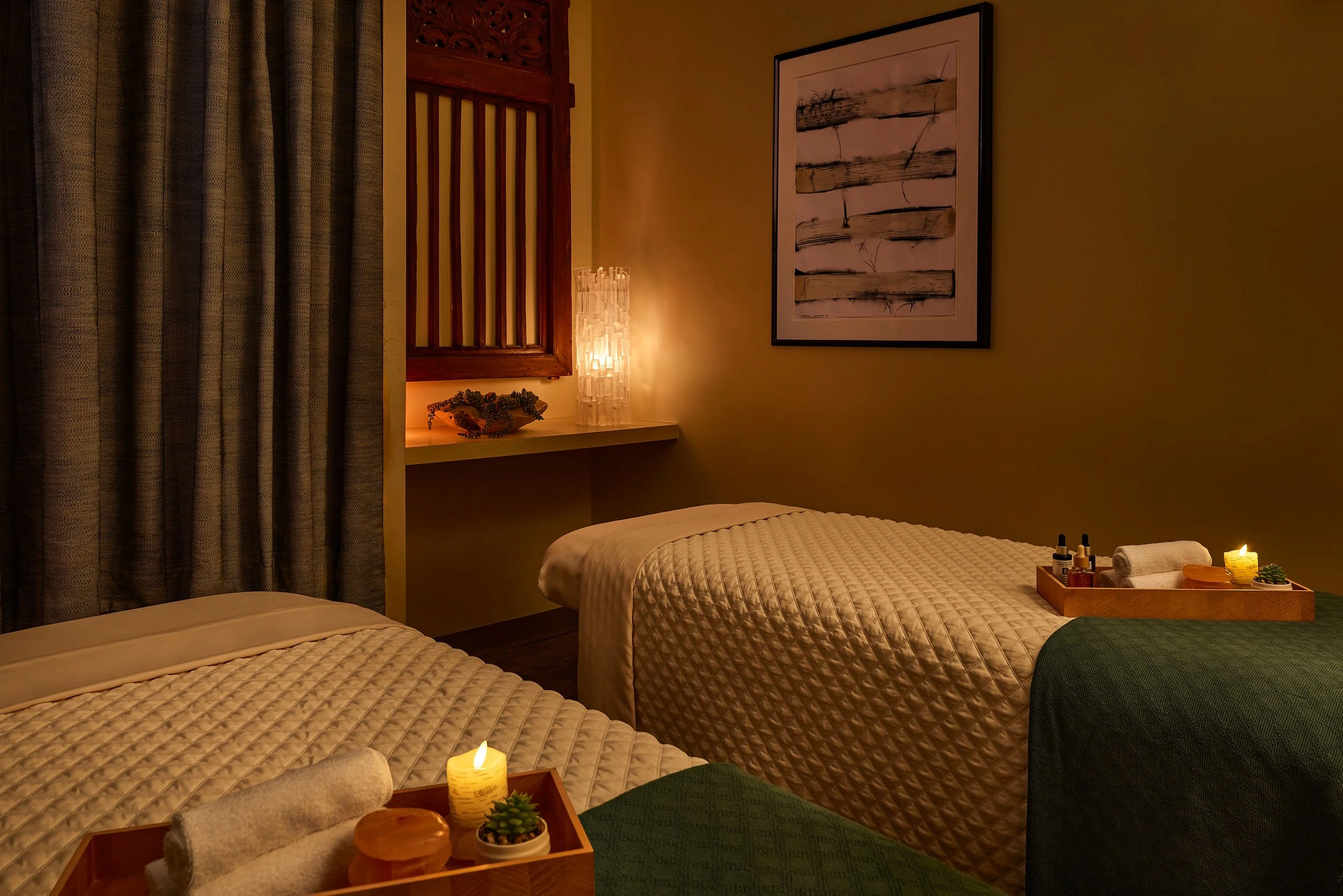 A cozy massage room with two massage tables covered in beige quilted blankets, a wooden tray with rolled towels, small bottles, a candle, and a small succulent plant. There are curtains on the left and artwork on the right wall, with a soft, warm lig