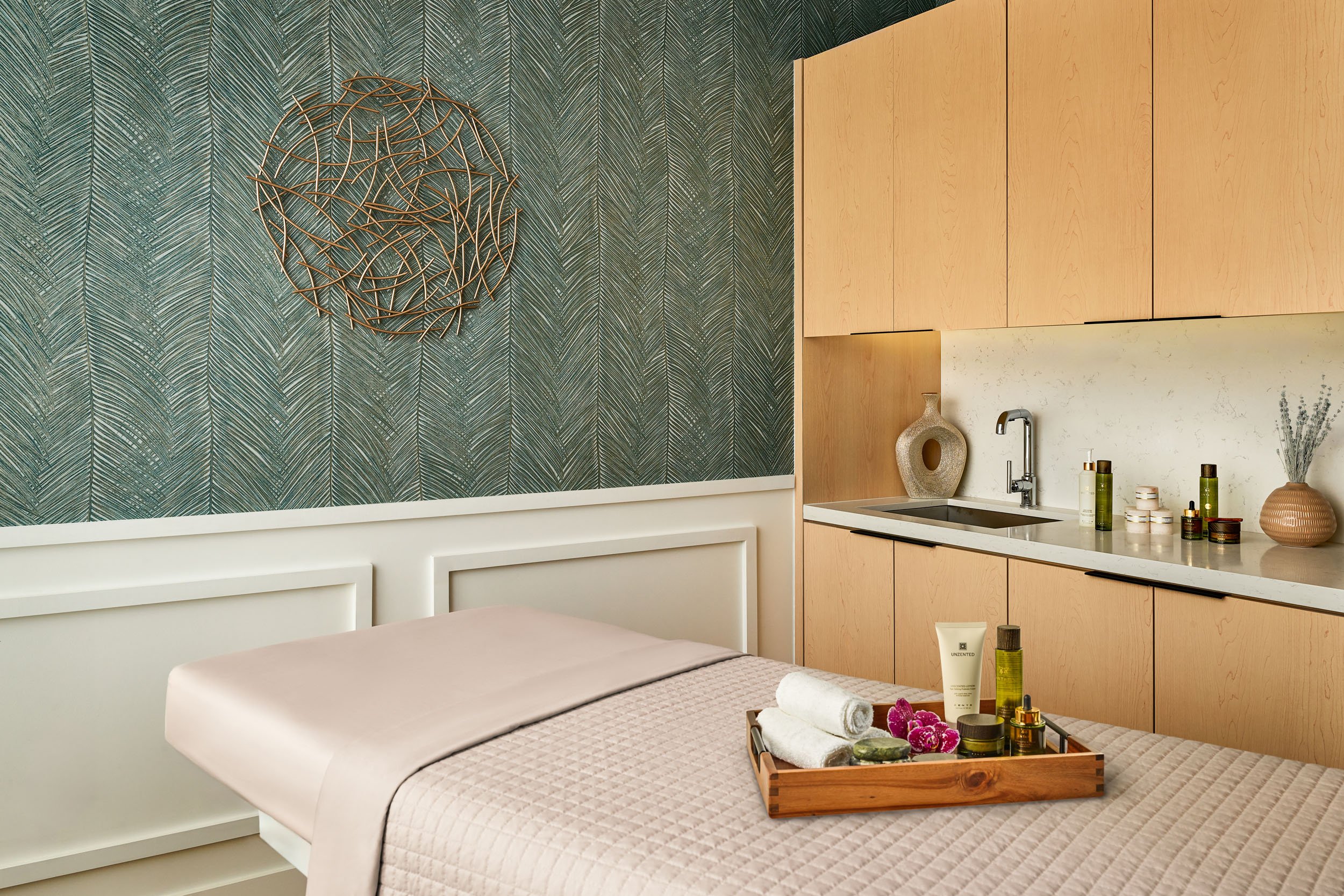 A bedroom with tropical leaf patterned wallpaper, a decorative wall sculpture, a white bed with a beige quilt, a small kitchen area with wooden cabinets, a sink, various skincare and beauty products on the counter, and a tray with towels and flowers 