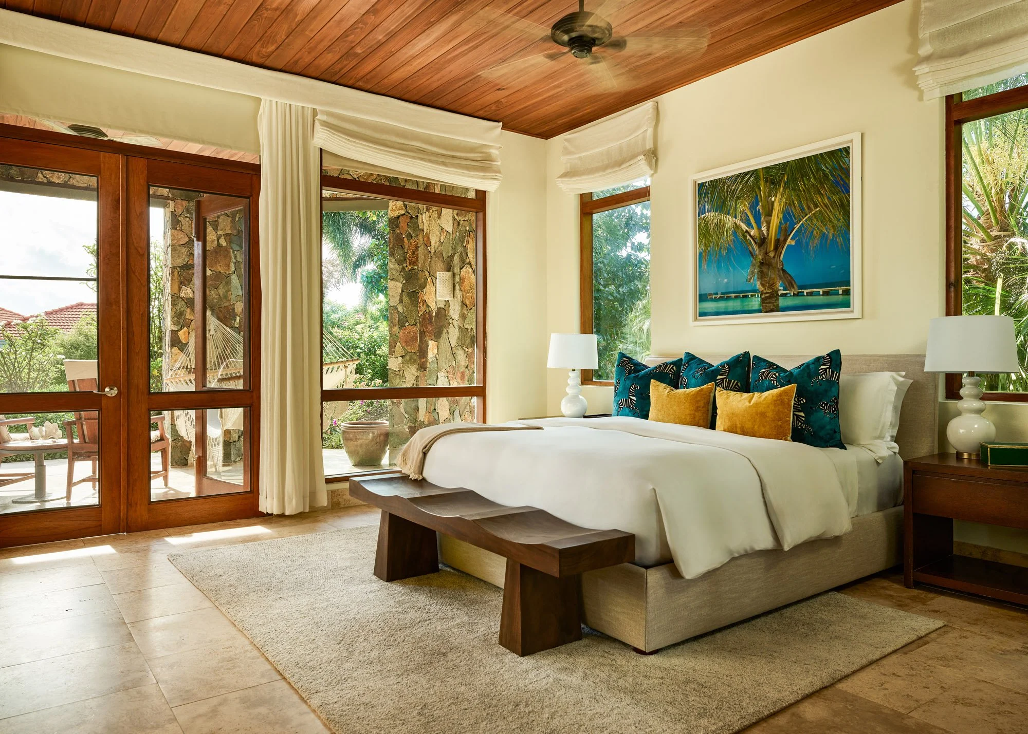 A bedroom with a large bed, colorful pillows, a nightstand with lamps on both sides, and large windows with white curtains showing greenery outside.