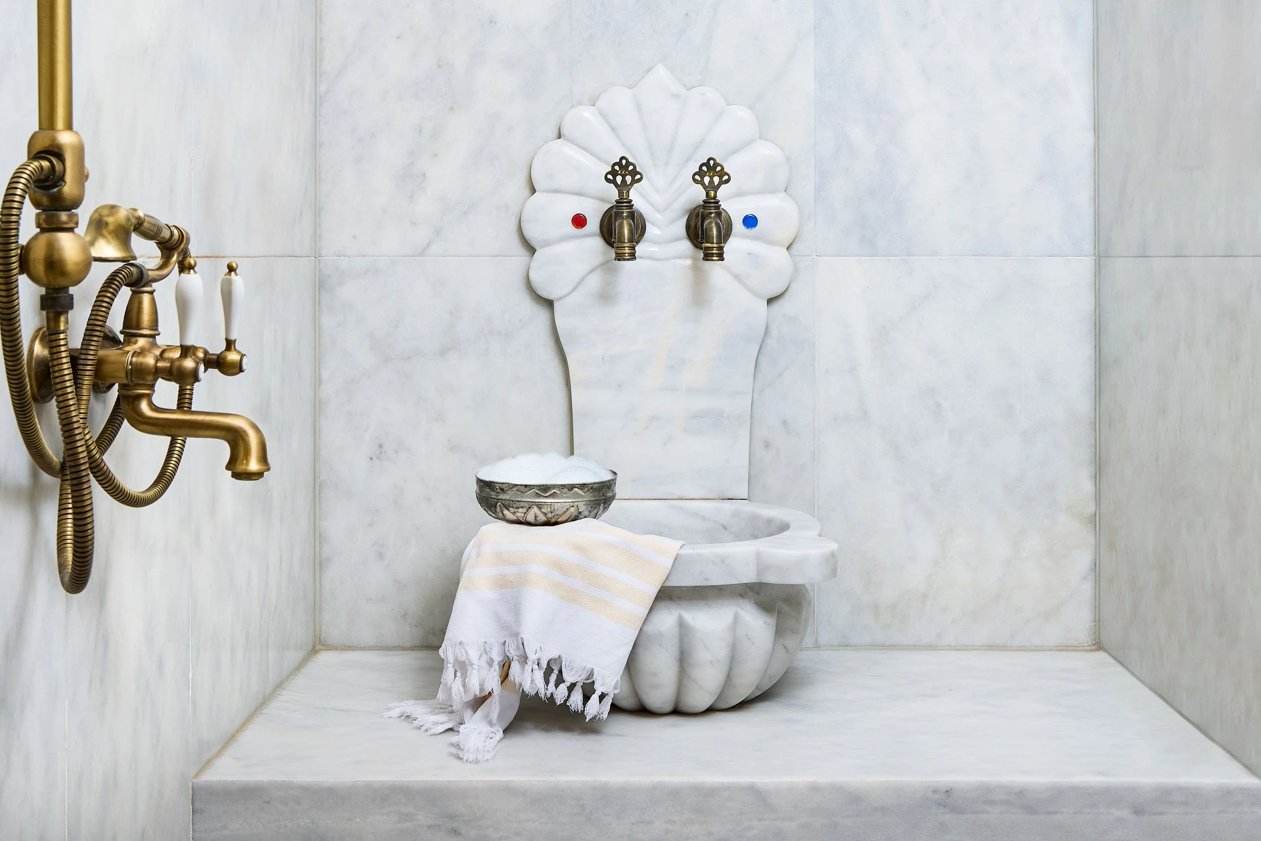 A restroom with a marble wall and floor, featuring a vintage-style toilet with a scalloped design, topped with a towel, and a flower-shaped marble backplate with two brass faucets marked with red and blue for hot and cold water, and a brass shower sy