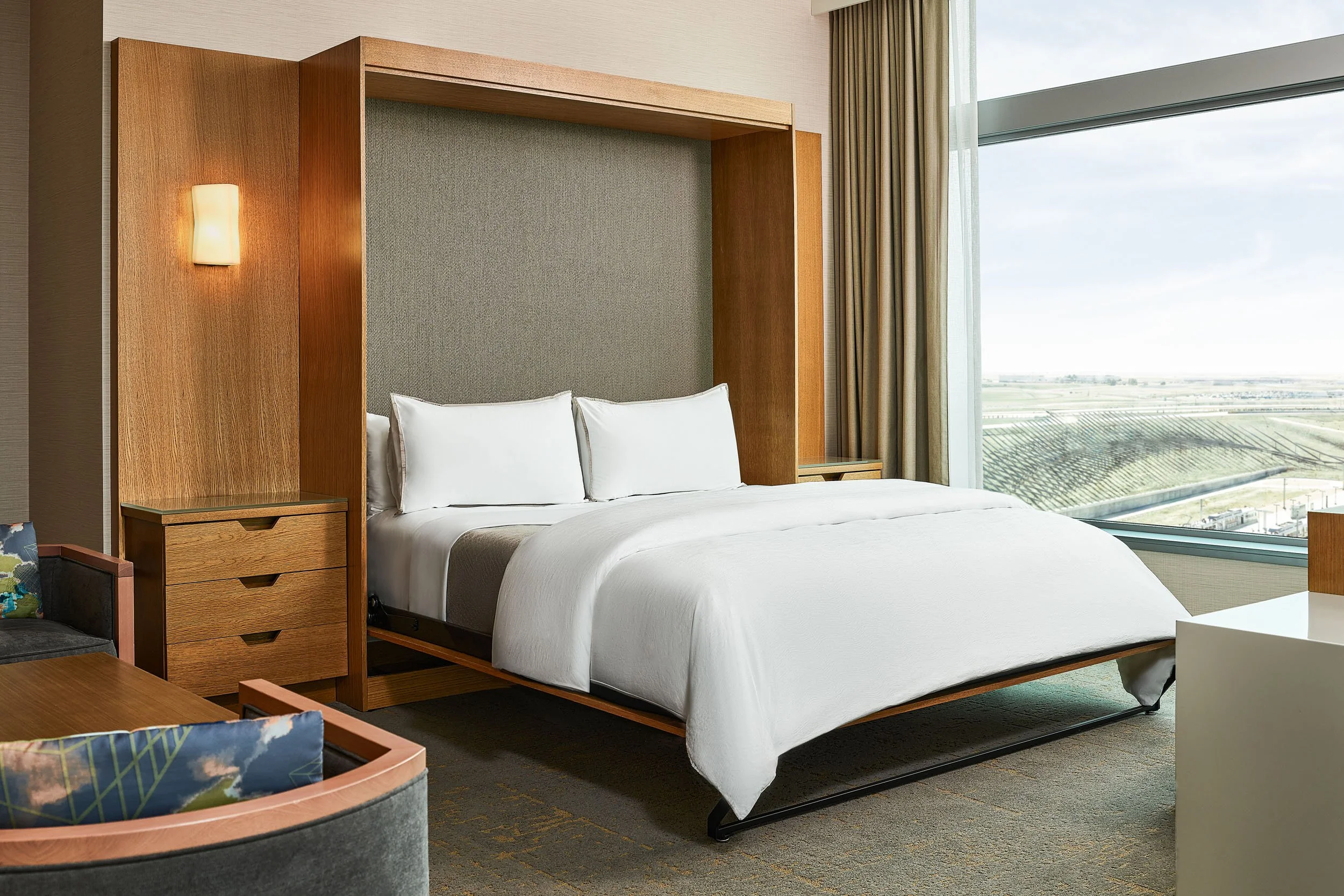A hotel room with a bed, wooden headboard, and large window showing a landscape view. The room has modern decor and a warm color palette.