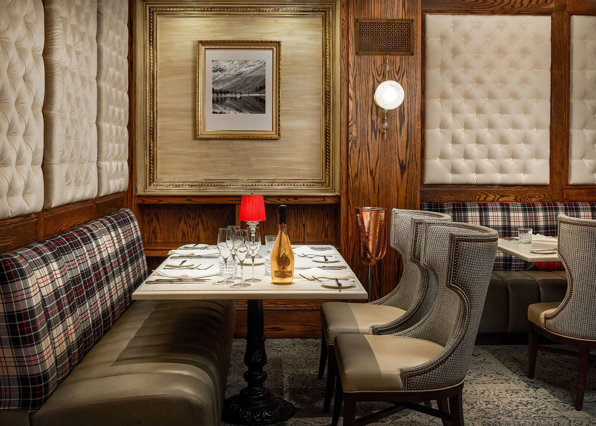 A cozy restaurant dining area with a mix of upholstered and plaid-patterned chairs around a white table set with wine glasses and tableware. The walls are decorated with tufted padded panels, framed artwork of a mountain landscape, and a spherical wa