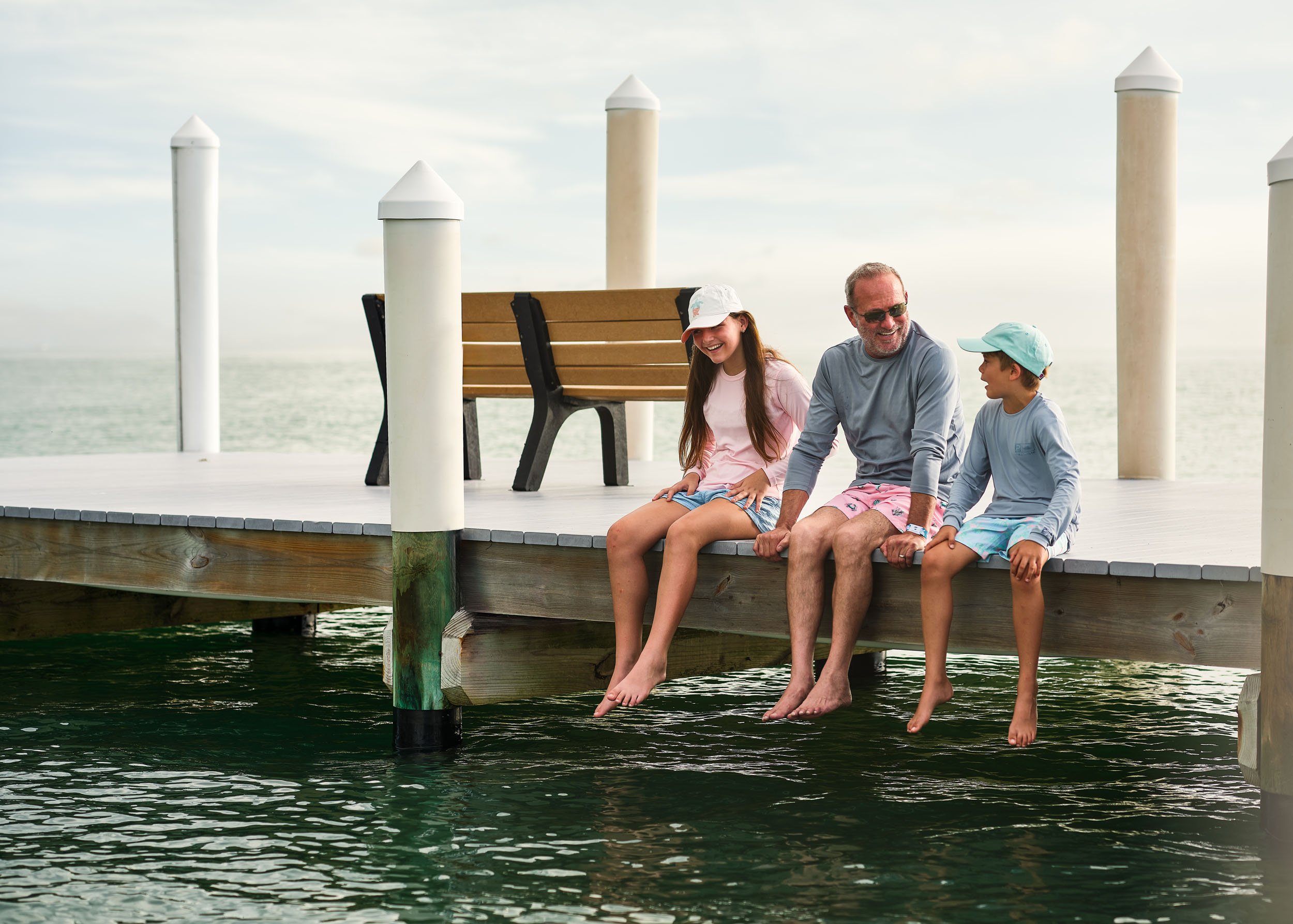 A family of three sitting on a dock with their feet dangling in the water, smiling and enjoying a day by the ocean.