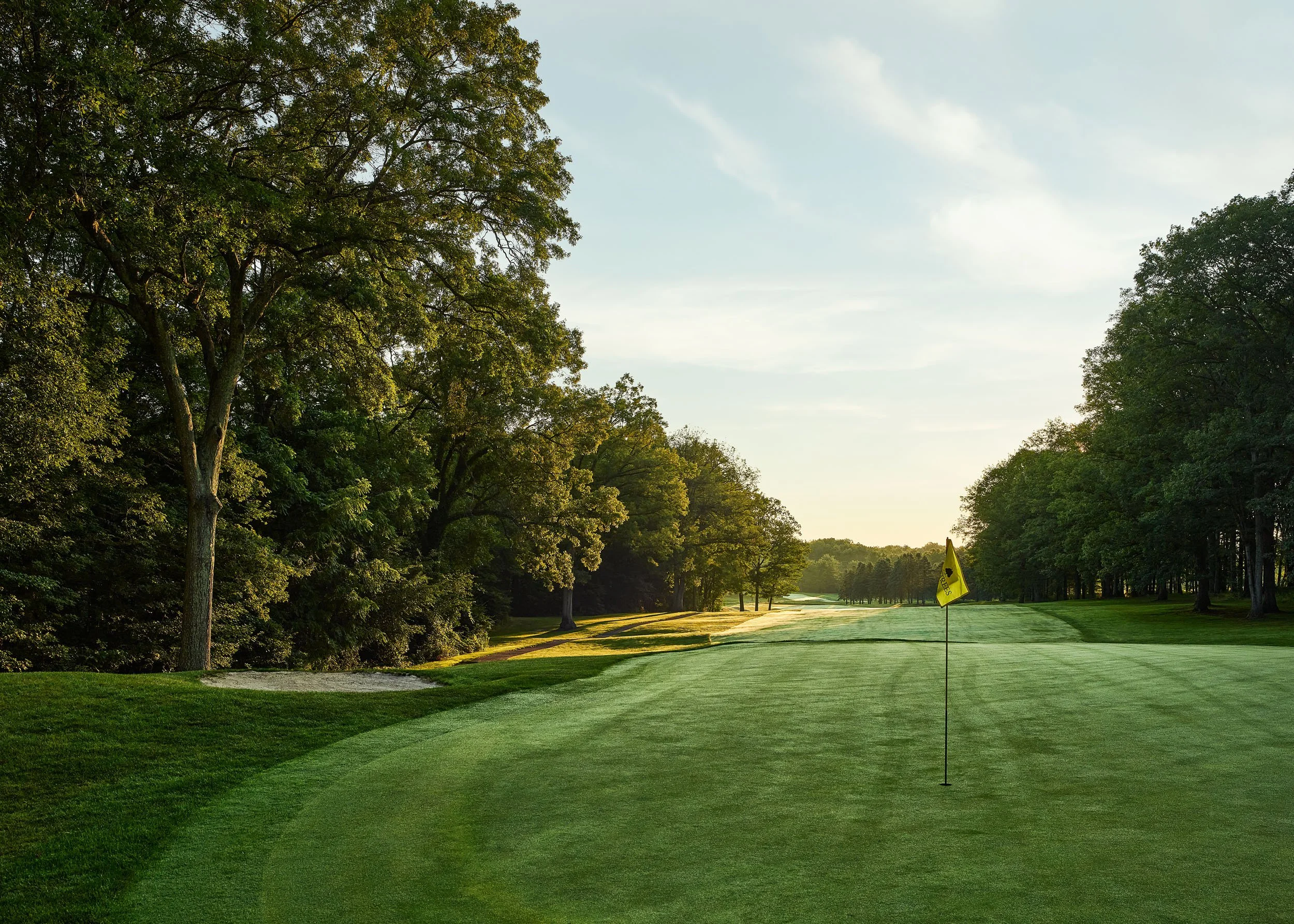 A golf course with a hole marked as number 3, surrounded by green grass and tall trees, under a partly cloudy sky during the daytime.
