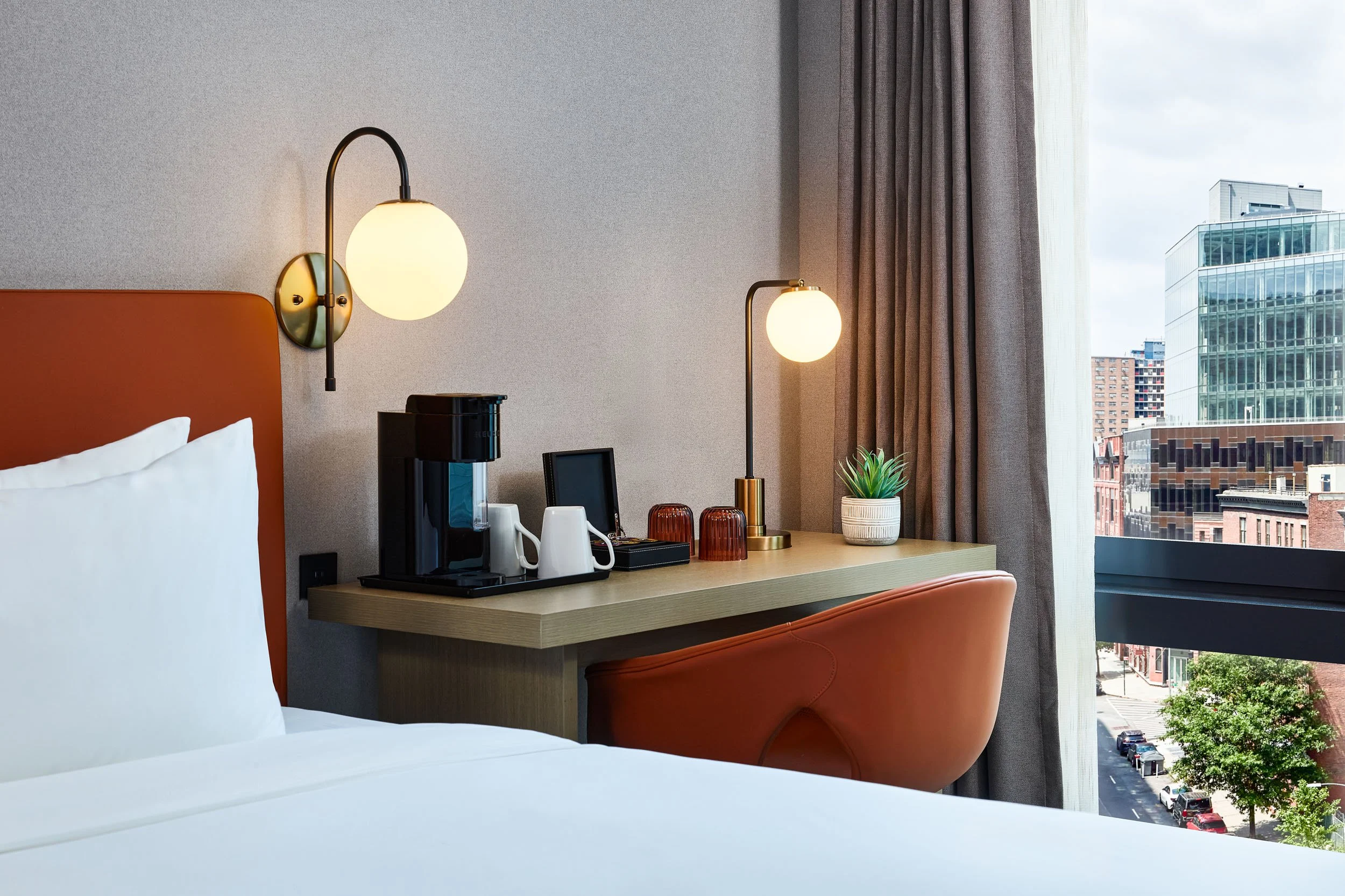 View of a hotel room with a bed, wall-mounted lamps, a countertop with a coffee maker, cups, and a plant. A large window shows a cityscape with modern glass buildings and a street below.