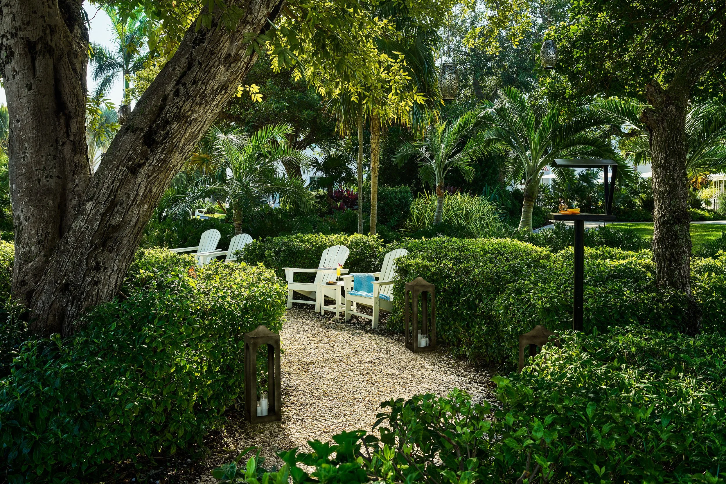 A lush garden with green trees and shrubs, white chairs along a gravel pathway, and decorative lanterns hanging from trees.