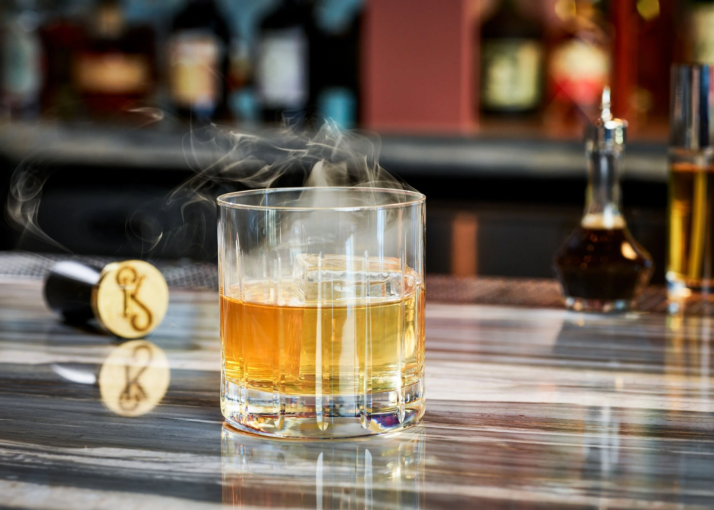 A glass filled with an amber-colored liquid, possibly whiskey, on a reflective surface with steam rising from it. In the background, there are blurred bottles on a shelf, and a small bottle with a dark liquid is visible on the right. A cork with a lo