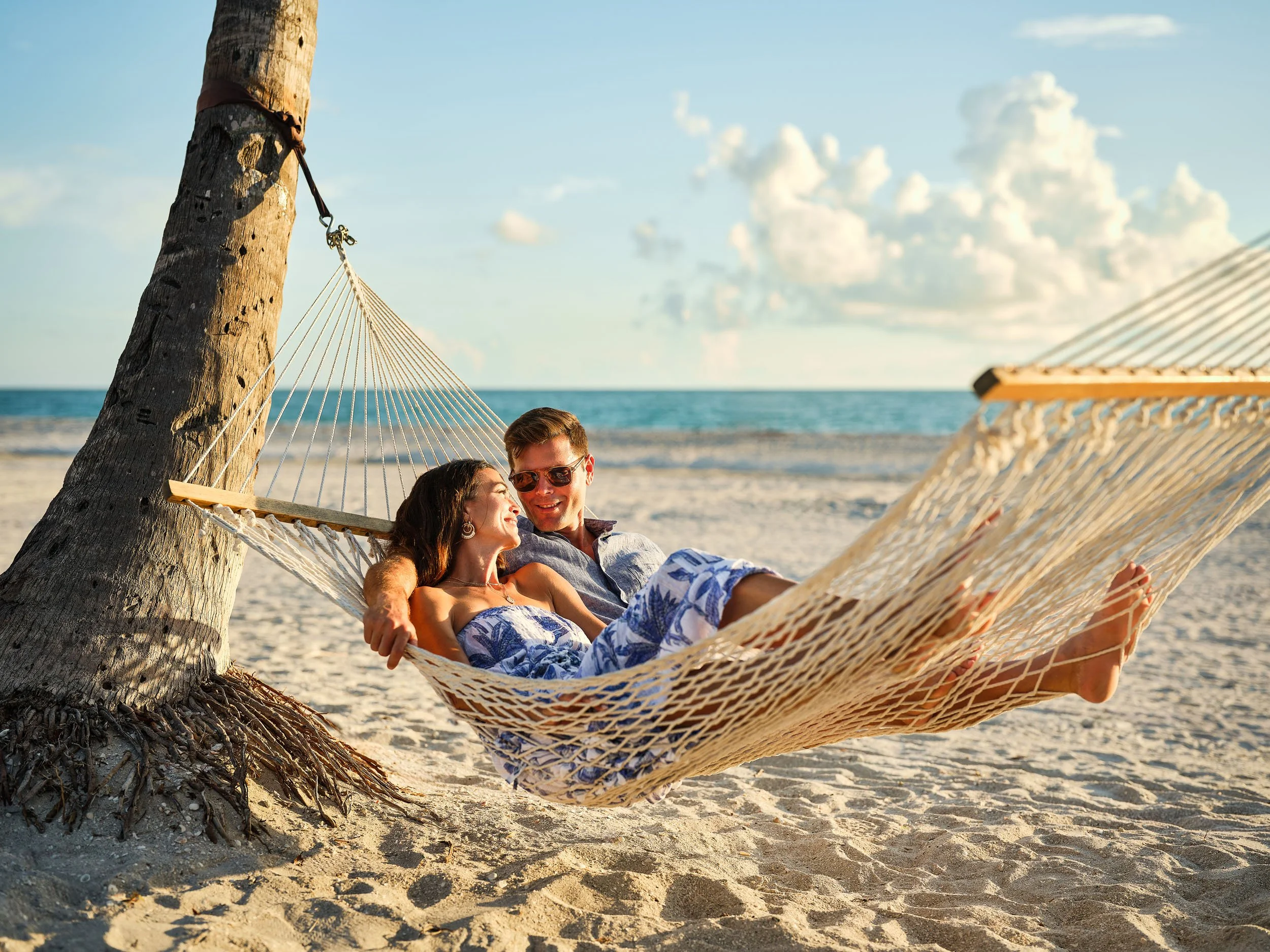 A couple relaxing on a hammock tied to a palm tree on a sandy beach with the ocean and clouds in the background during sunset.