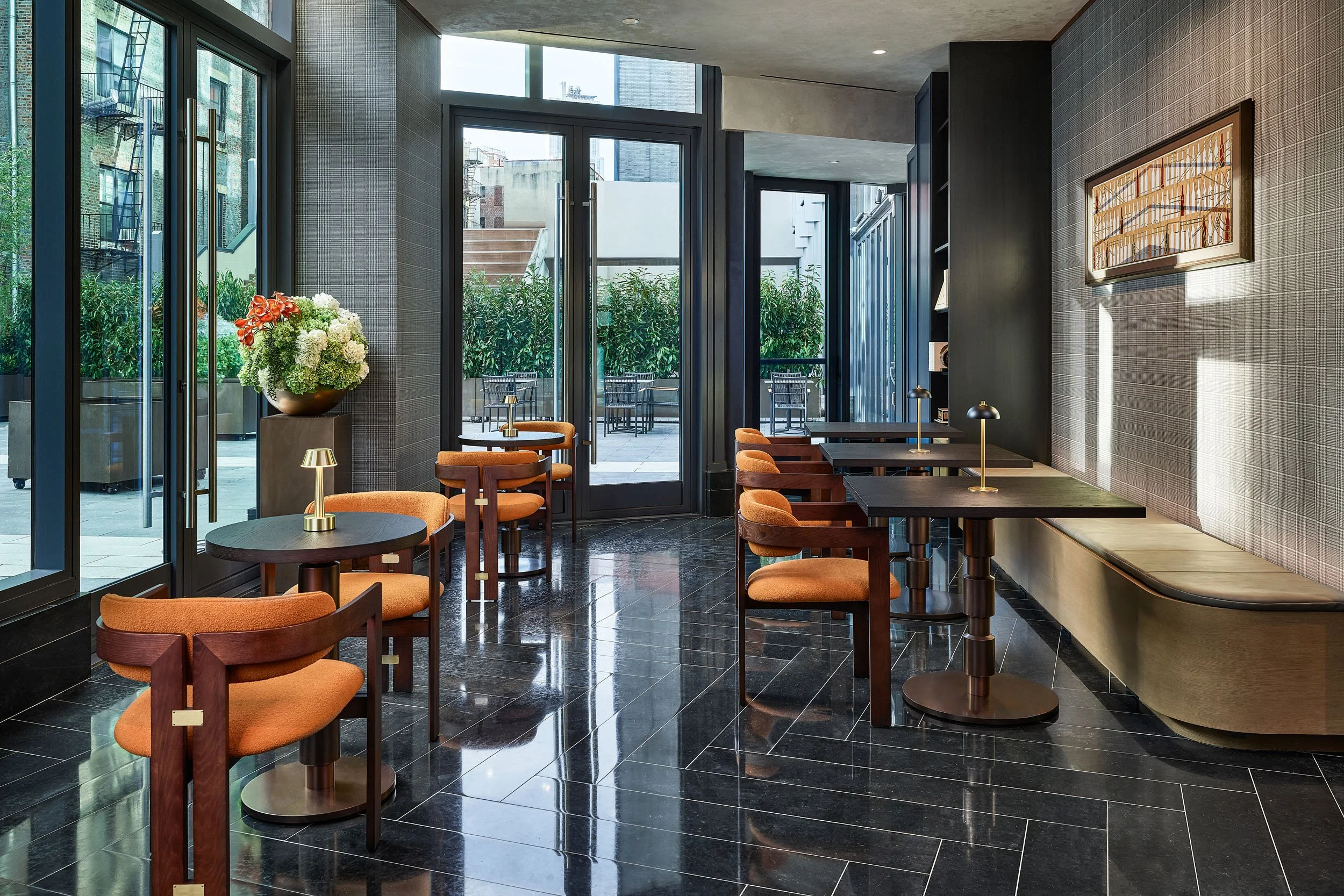 Interior of a modern cafe or restaurant with black tile flooring, dark walls, wooden furniture with orange upholstered seats, large glass doors and windows, and outdoor seating visible outside. There is a large floral arrangement in a vase near the w