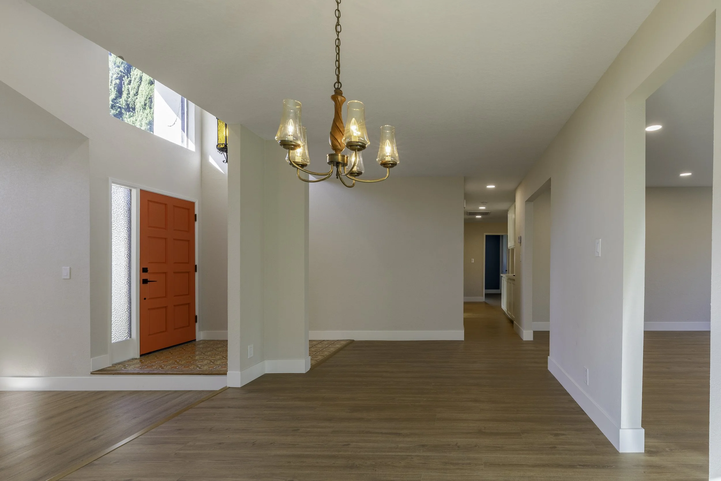 Entryway of a house with a red front door, a chandelier hanging from the ceiling, and a split-level floor with hardwood and tile sections. Large windows let in natural light.