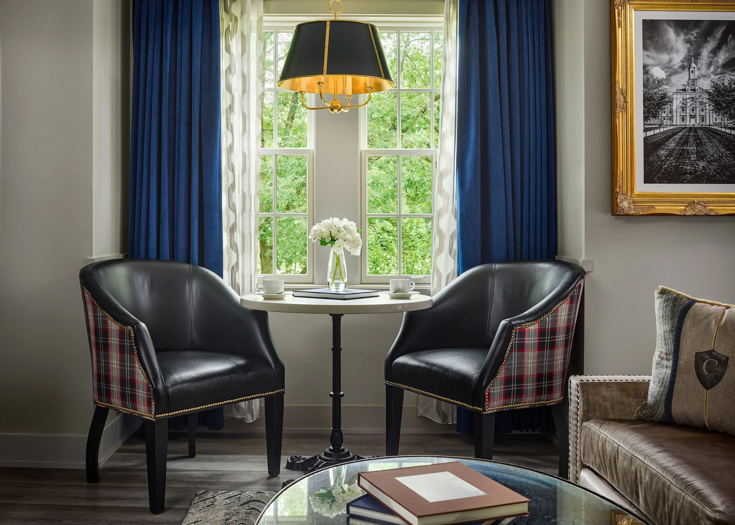 A round dining table with a white top, set with a clear vase of white flowers and a few books, surrounded by four black leather chairs with plaid fabric on the back, in front of a window with blue and white curtains and a black and gold chandelier ha