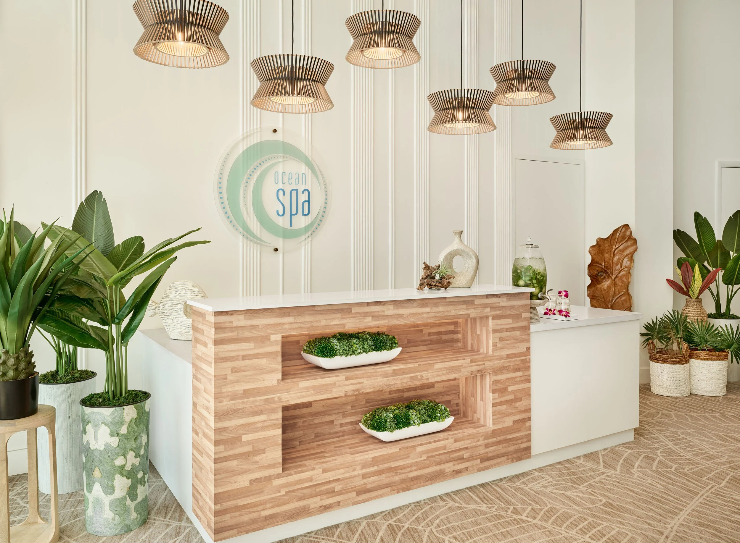 Interior of a spa with a reception desk, green plants, and decorative vases under modern hanging lights.