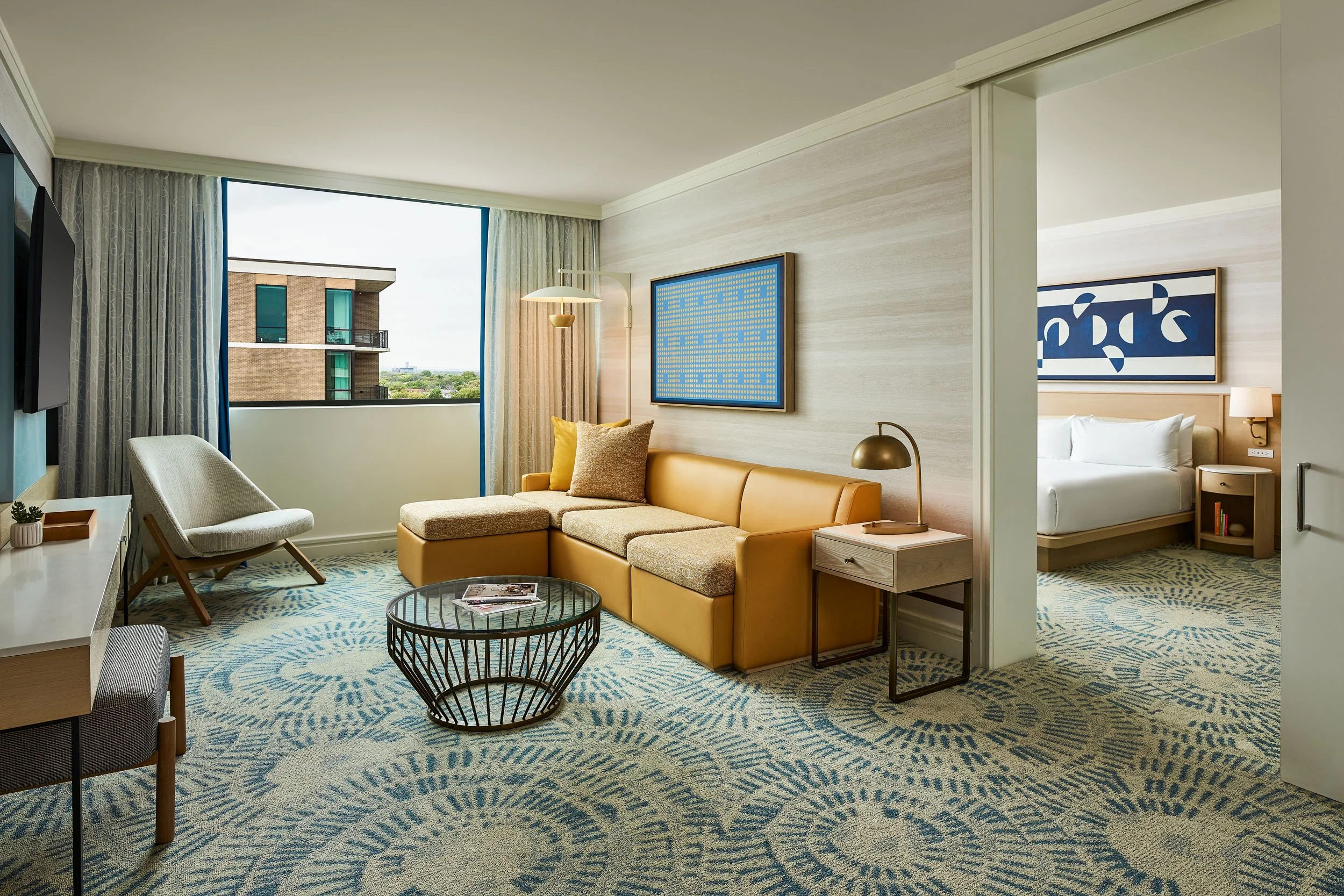 Living room with large window, beige sofa, patterned blue rug, framed artwork, white chair, small round coffee table, and a bedroom with a bed and nightstand visible through an open doorway.
