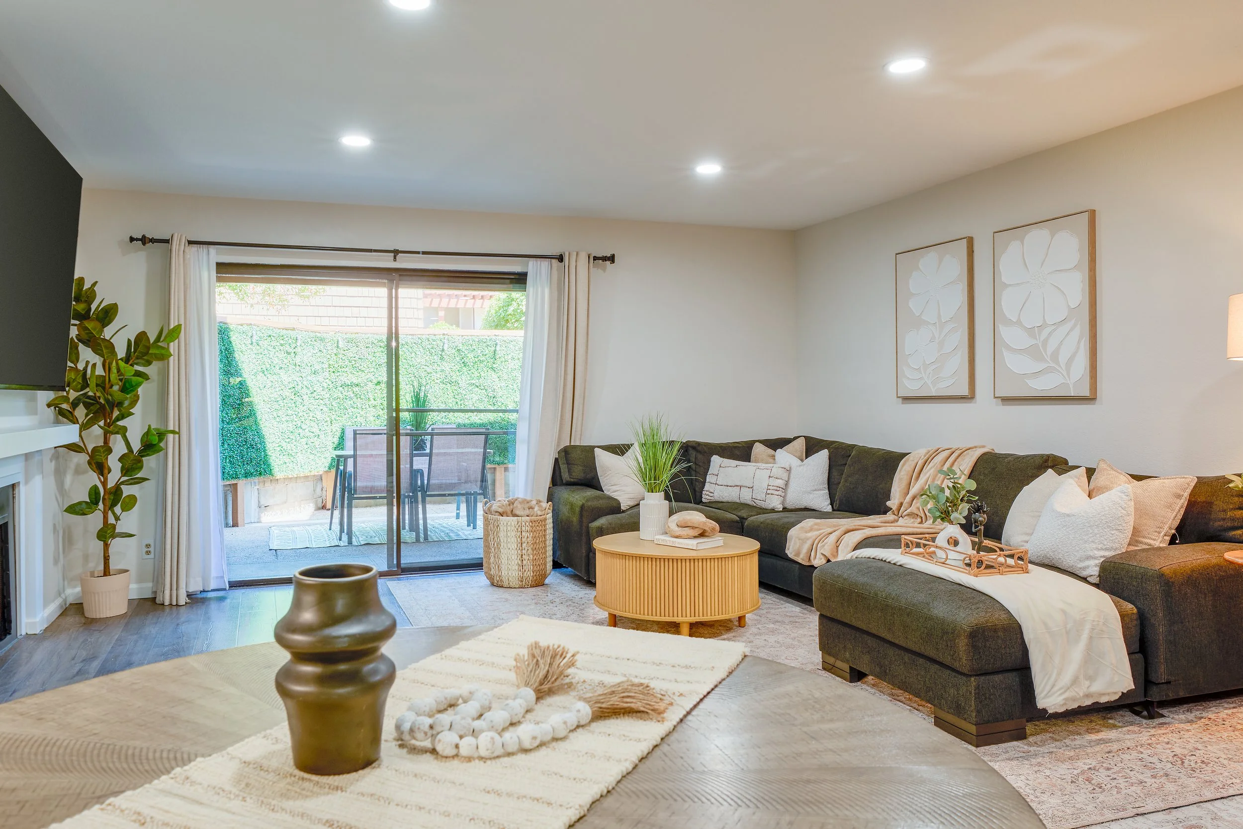 Living room with dark sectional sofa, beige and patterned pillows, light wood coffee table, and wall art of white flowers. There is a sliding glass door leading to an outdoor patio with dining table and chairs, surrounded by greenery. Indoor plants a