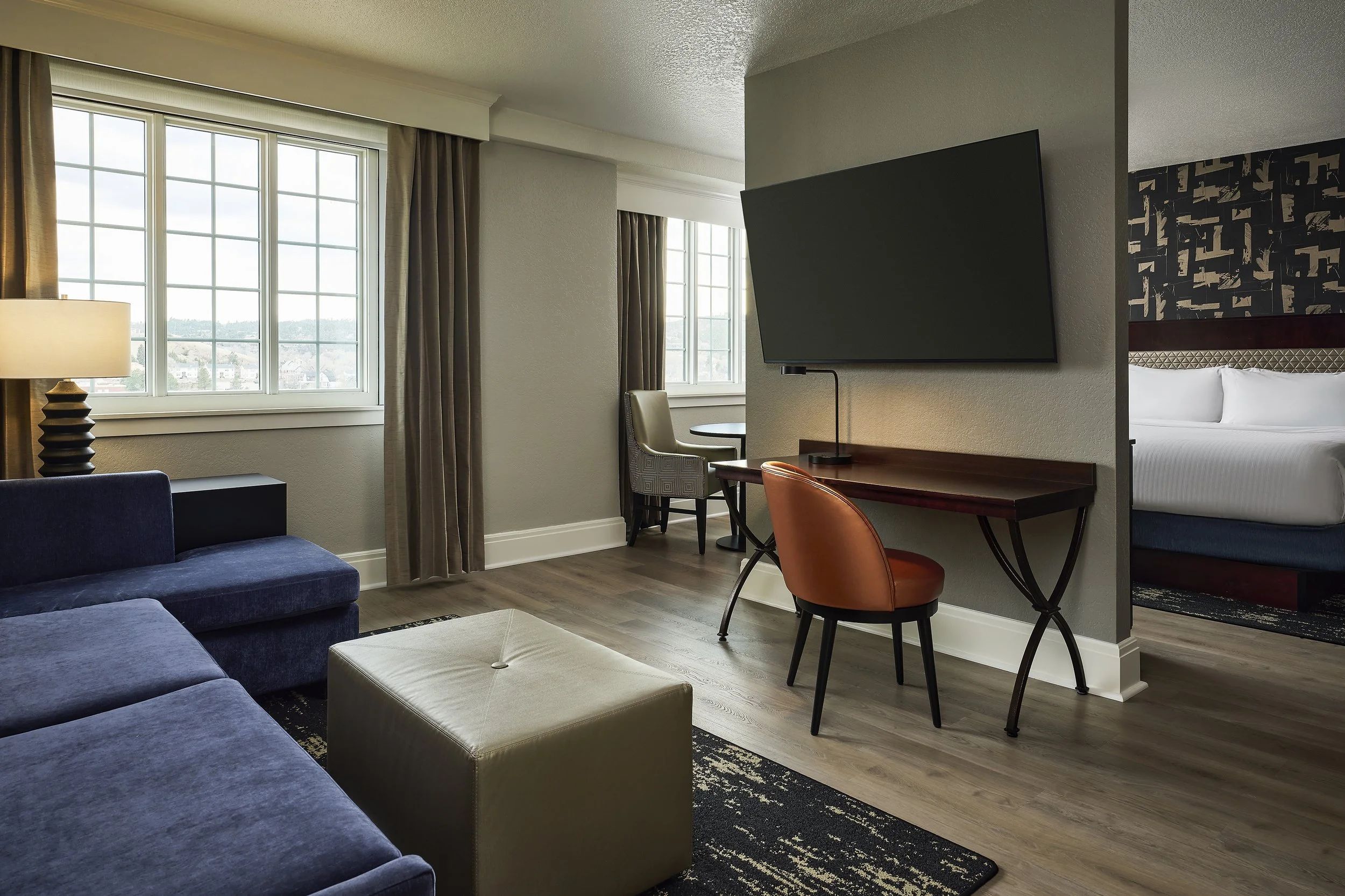 Interior of a modern hotel room with large windows, a blue sofa, a beige ottoman, a wall-mounted flat-screen TV, a small round table with two chairs, and a bed with white linens visible in the background.
