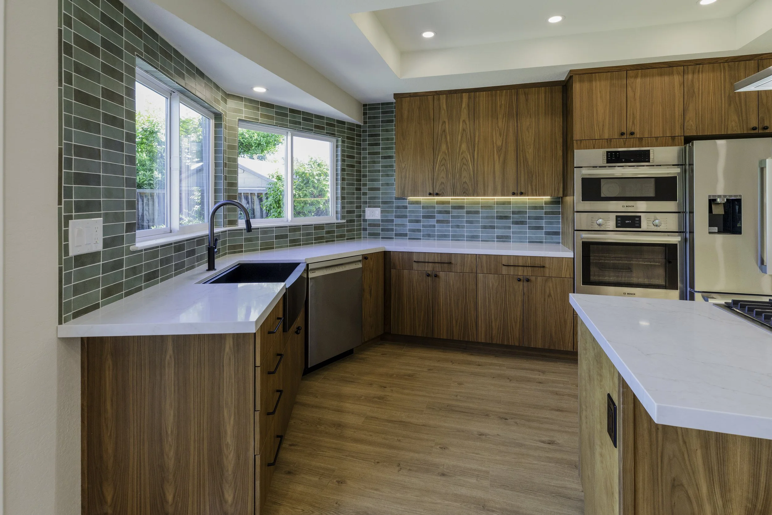 Modern kitchen with green tile backsplash, wooden cabinets, stainless steel appliances, and a window showing greenery outside.