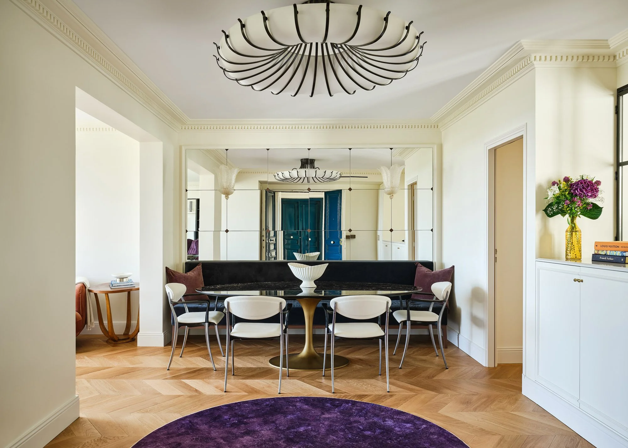 A modern dining room with a round black marble table surrounded by white and black chairs. A gold pedestal base supports the table. There is a large purple rug beneath the table. The walls are painted a light cream color, and a mirror wall reflects t