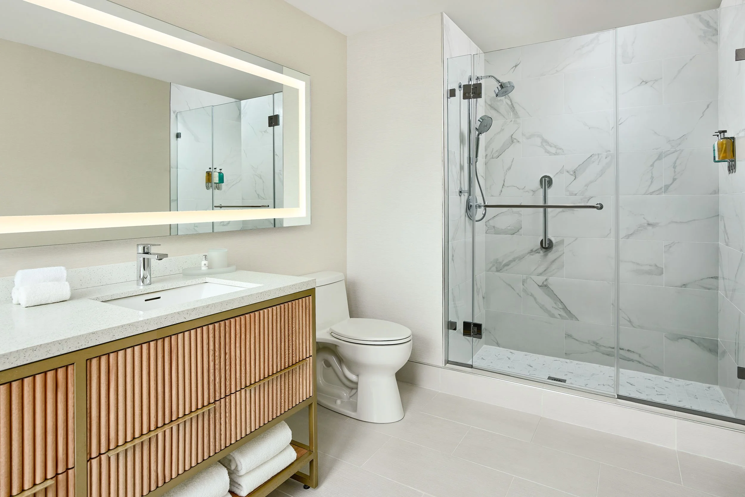 Modern bathroom with a large illuminated mirror, a white sink, a cabinet with vertical wooden slats, a toilet, and a glass-enclosed shower with marble walls and a grab bar.
