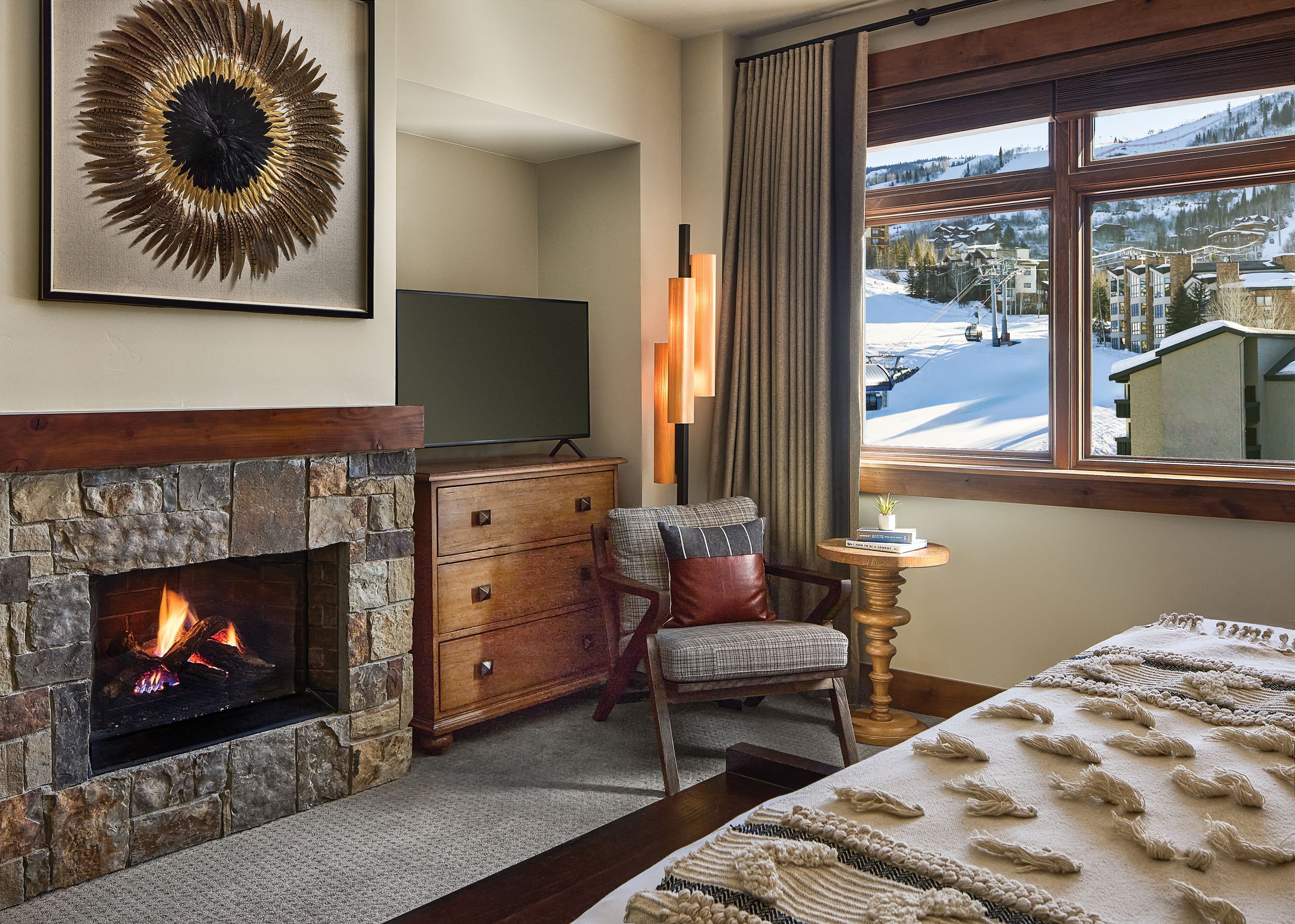 A cozy living room featuring a stone fireplace with a small fire burning, a wooden chest of drawers, a plaid armchair, a small wooden side table with a plant and books, a bed with a textured throw blanket, large windows with a snowy mountain landscap