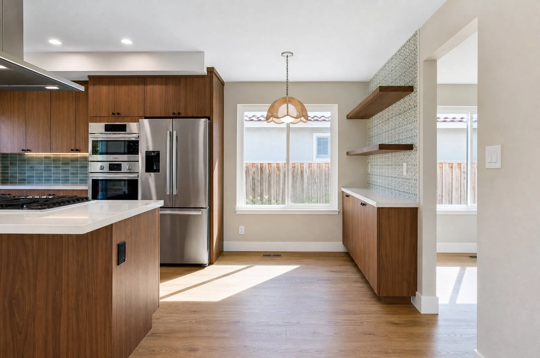 Modern kitchen with wooden cabinets, stainless steel refrigerator, green tile backsplash, and a beige patterned accent wall with two wooden shelves, illuminated by natural light through a window.